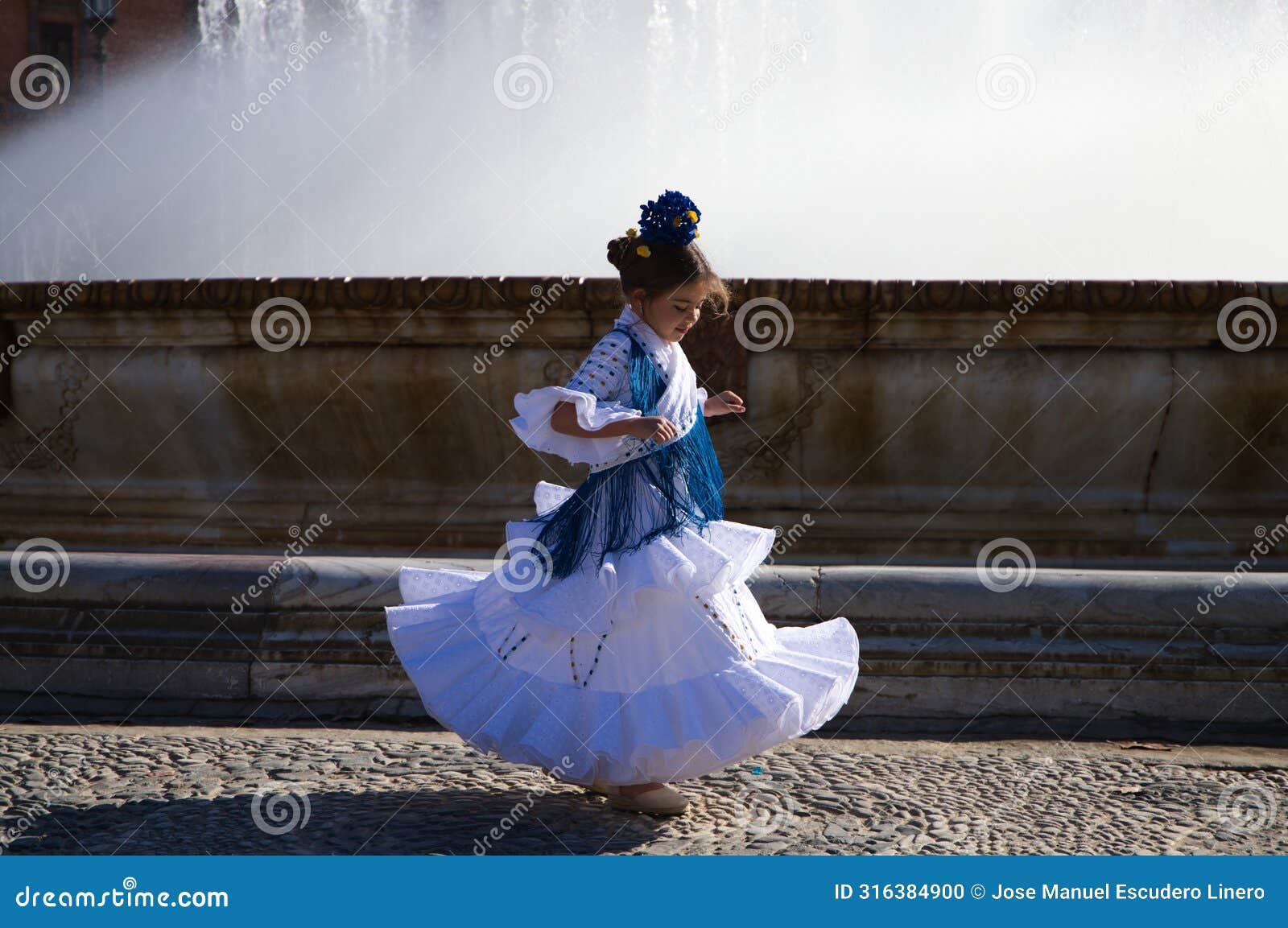 a pretty little girl dancing flamenco dressed in a white dress with ruffles and blue fringes in a famous square in seville, spain