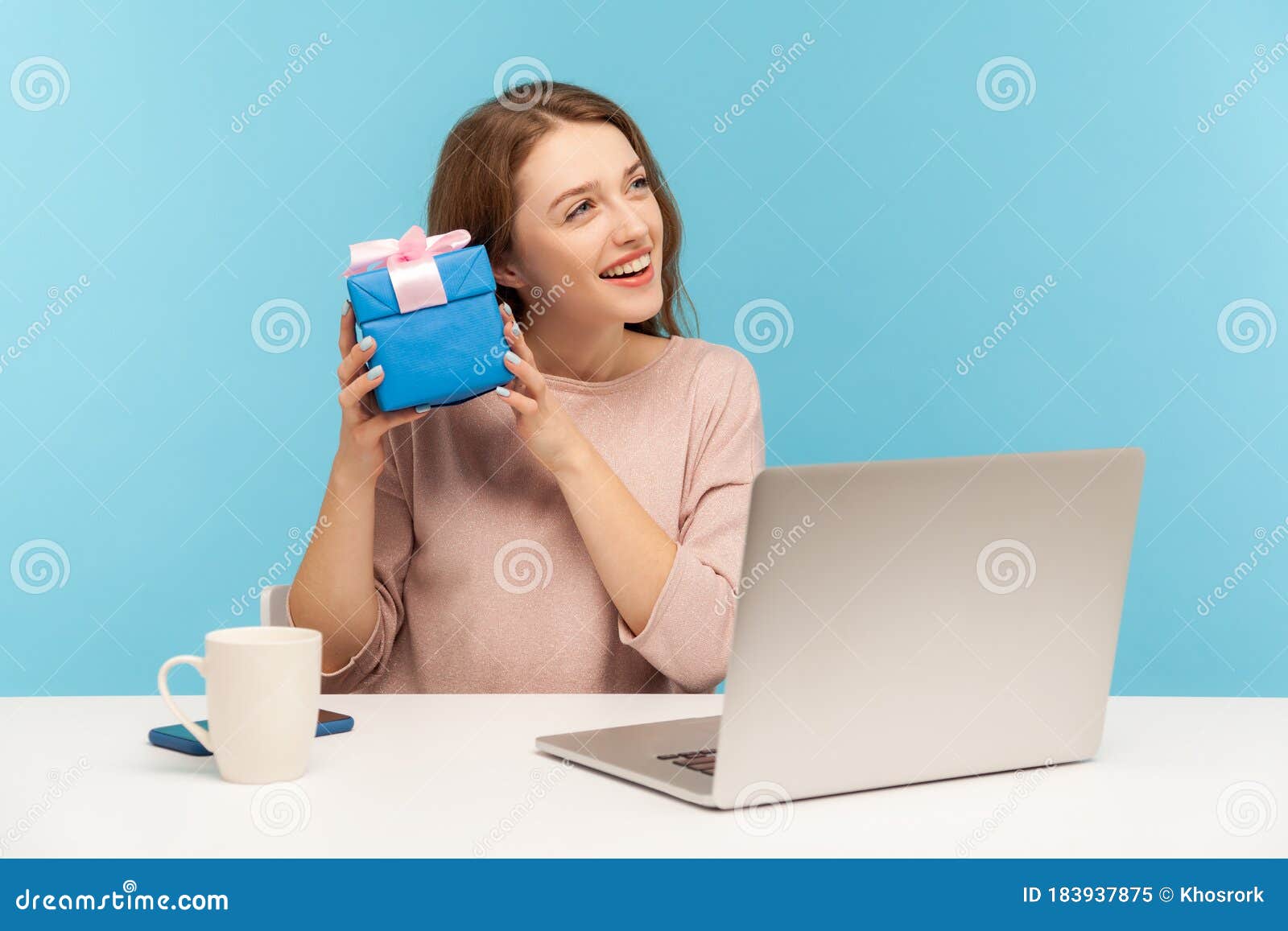 pretty woman employee sitting at workplace and holding gift near ear, listening with interest what`s inside box