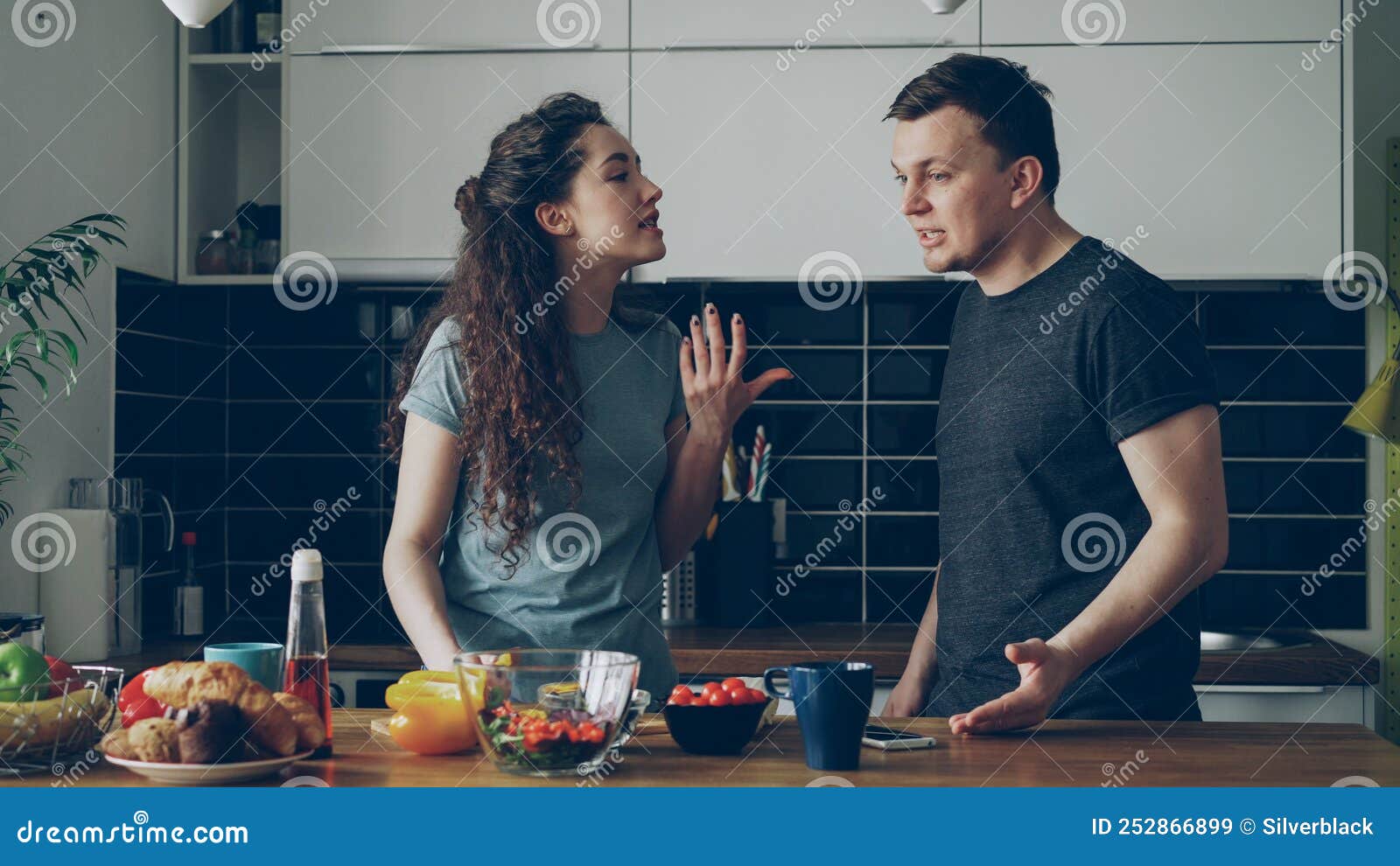 curly pretty woman standing at table cutting salad while her boyfriend comes and they argueing speaking loud and nervous