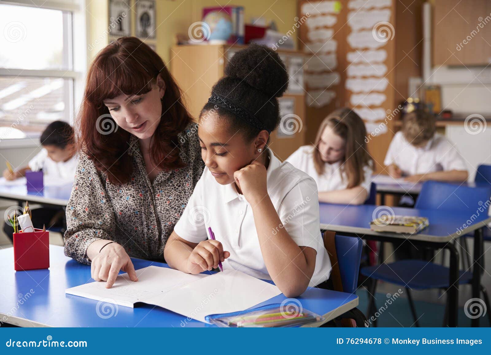 primary school teacher helping a schoolgirl at her desk