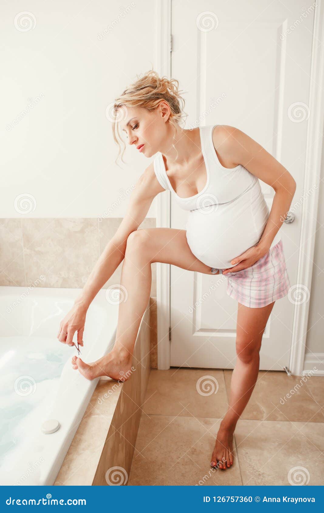 pregnant blonde caucasian woman cutting pare finger nails on legs toes in bathroom.