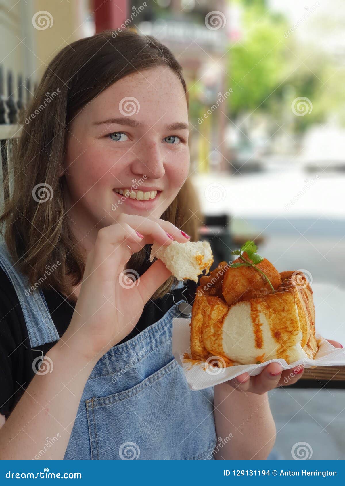 Profil der lächelnden Frau ungefähr, zum von Bunny Chow zu schmecken. Die junge lächelnde Frau, die ein Straßenlebensmittel Durbans Bunny Chow schmeckt, das ein südafrikanischer Schnellimbissteller ist, der aus einem ausgehöhlten heraus Brotlaib besteht, füllte mit Curry