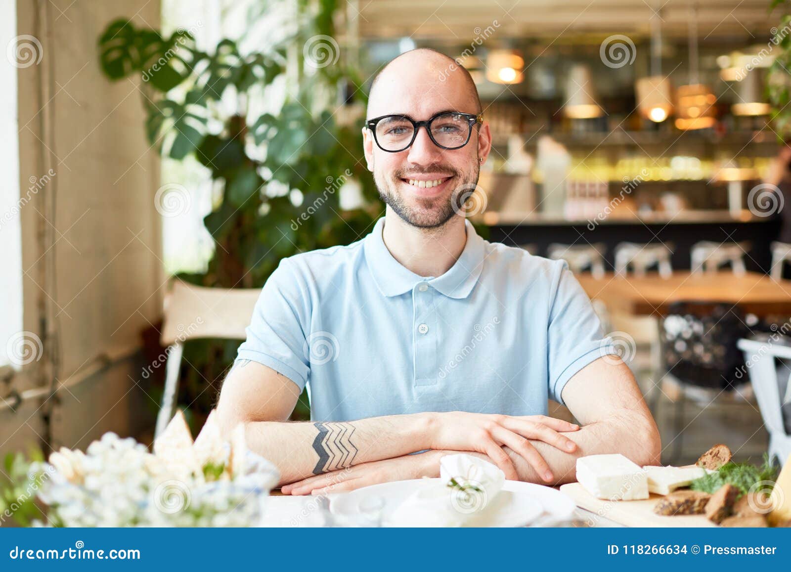 Quedese calvo al hombre barbudo que sonríe y que mira la cámara. Varón joven calvo hermoso en los vidrios que se sientan con las manos cruzadas y que sonríen en fondo del restaurante borroso