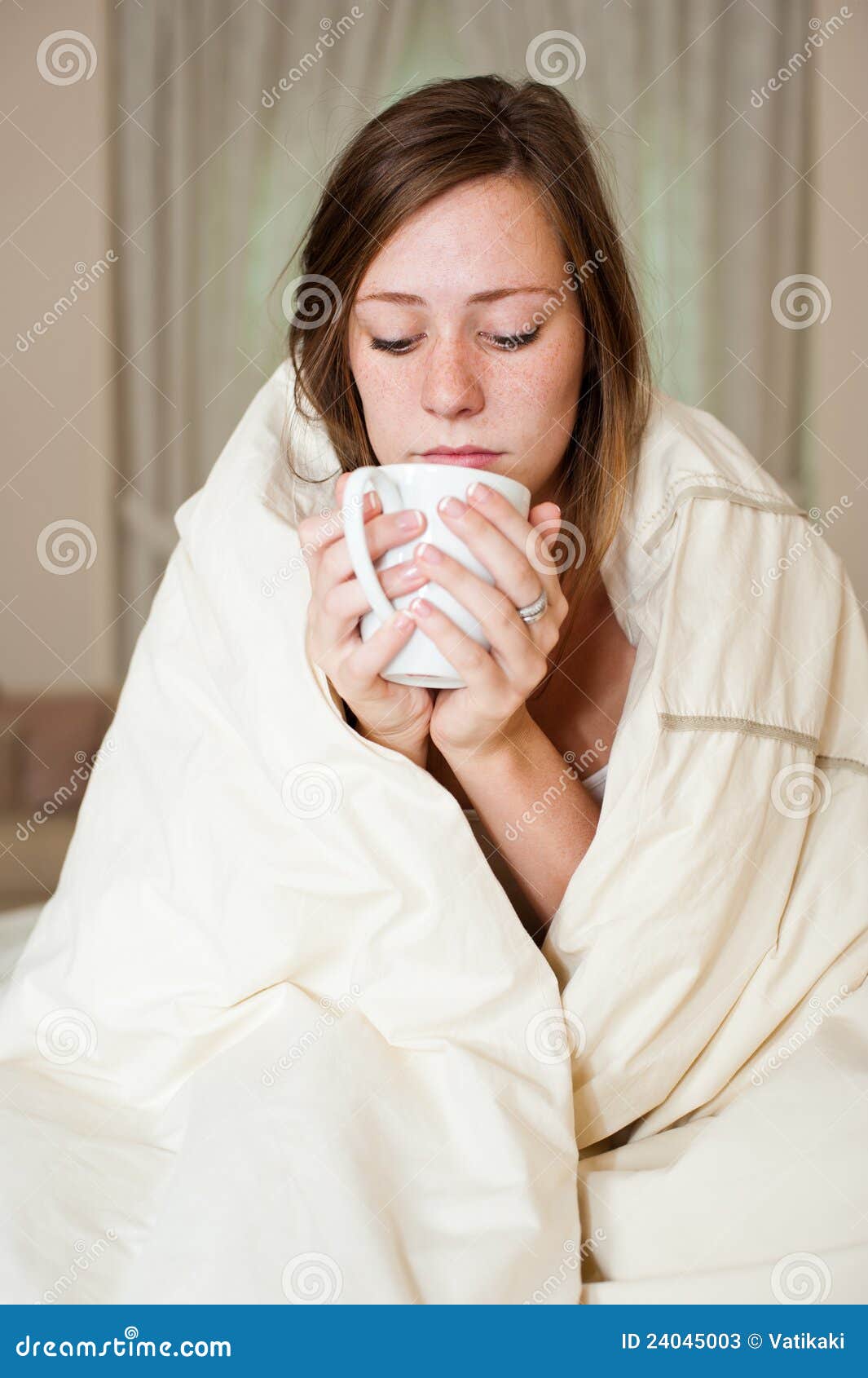 Ragazza ammalata. Una giovane signora ammalata in base, sedentesi con una coperta sopra le sue spalle e una tazza della bevanda calda.