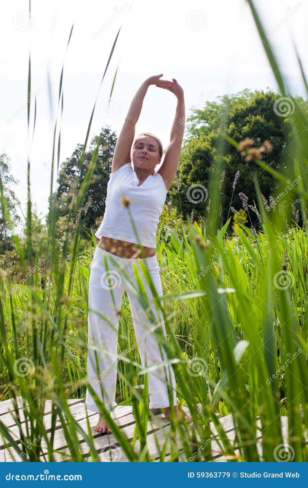 Ragazza bionda di zen 20s che si esercita nei surrondings verdi. All'aperto esercizio di zen - la giovane donna sorridente di yoga che outstretching le sue armi, chiudentesi osserva nella luce del giorno dell'estate, nella priorità alta verde del parco e nel fondo