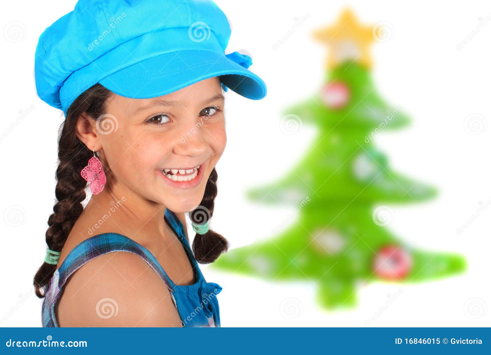 Ragazza di natale. Ragazza etnica adolescente graziosa di dieci anni una multi con le trecce ed albero di Natale nei precedenti che portano un cappello blu variopinto e un grande sorriso toothy
