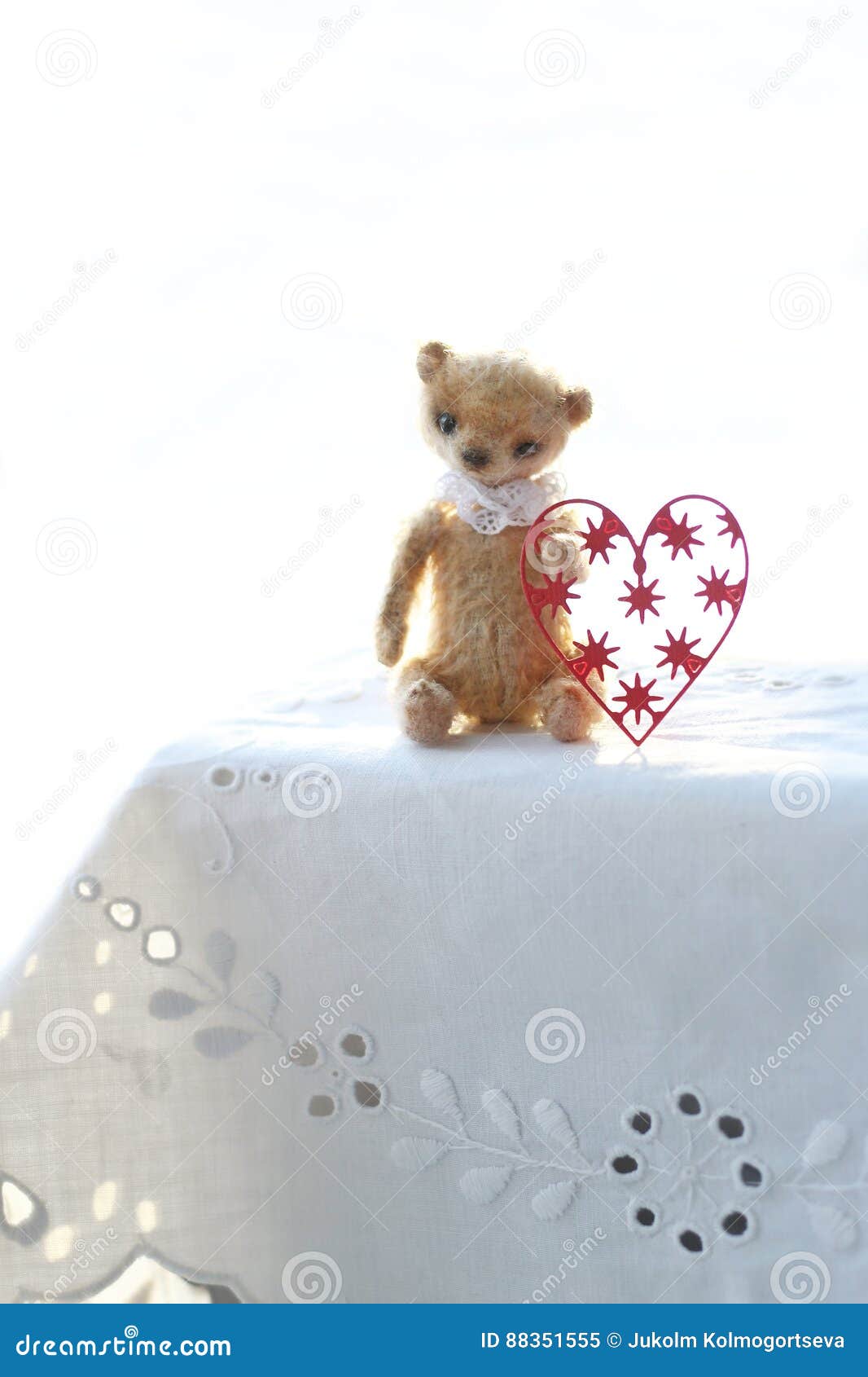 red paper heart and the small brown toy bear made of wool sitting on a white napkin warm colors. soft focus.