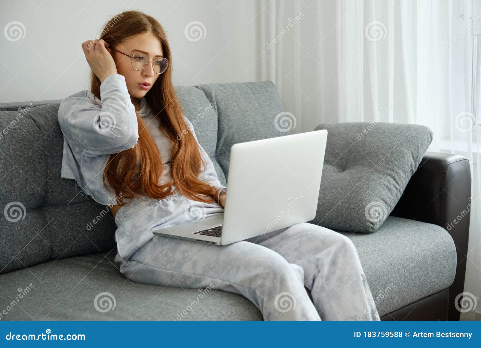 Redhead Girl In Pajamas And Glasses Sits On A Sofa With A Laptop