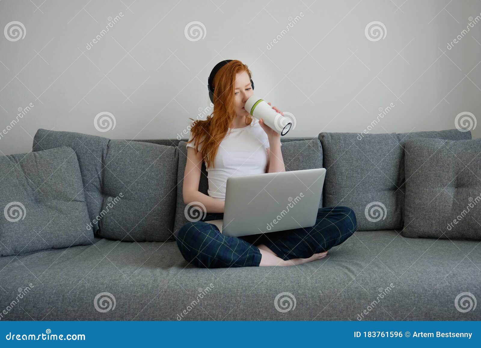 Redhead Girl In Pajamas Sits On A Sofa With A Laptop On Her Folded Legs