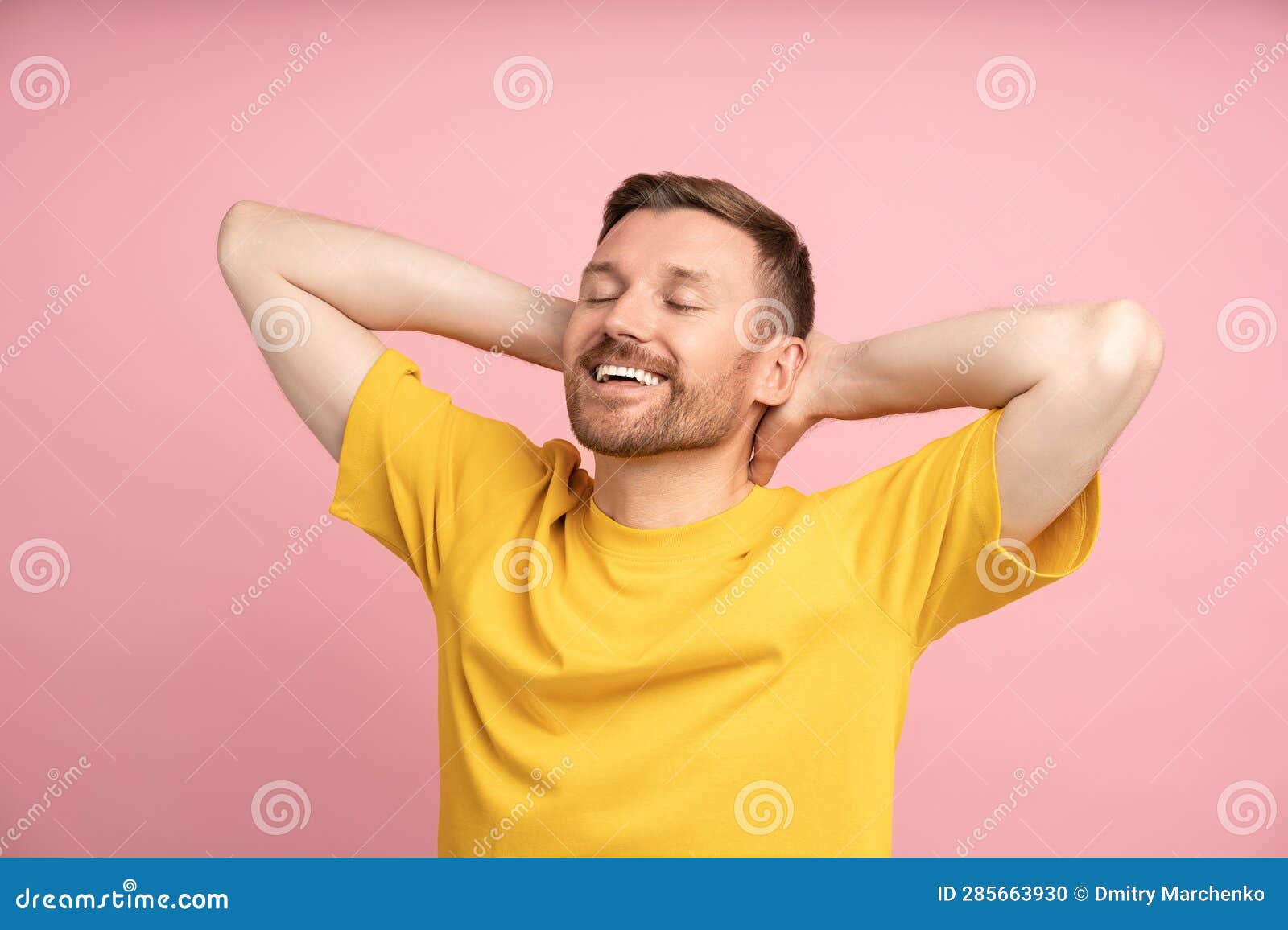 relaxed man holding hands behind head smiling with closed eyes standing on studio pink background.
