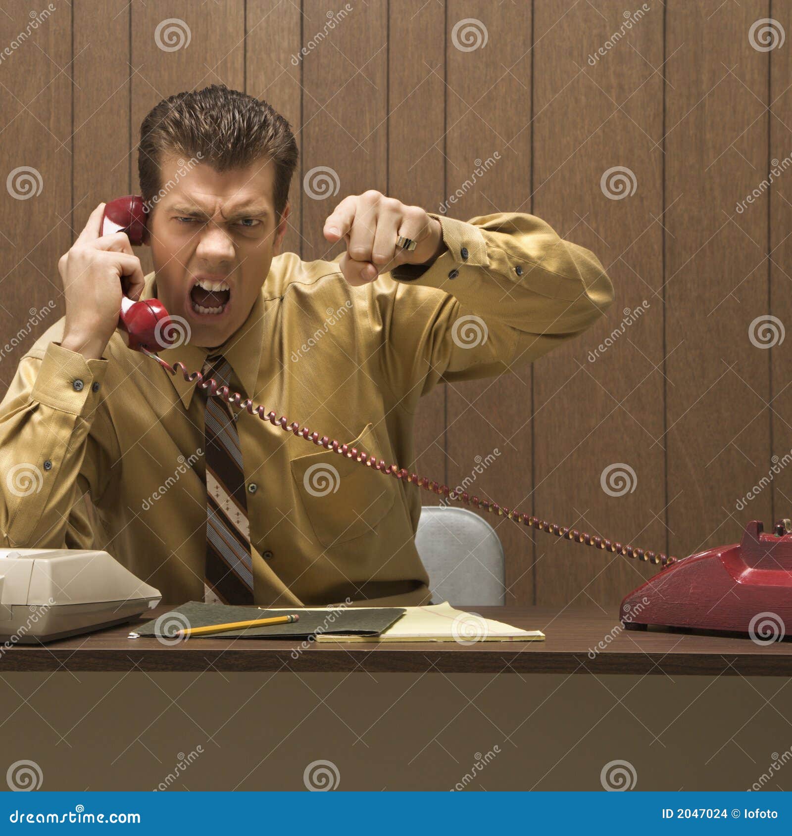 Retro business scene of angry man at desk. Caucasion mid-adult retro businessman sitting at desk talking on telephone with angry expression and gesture.