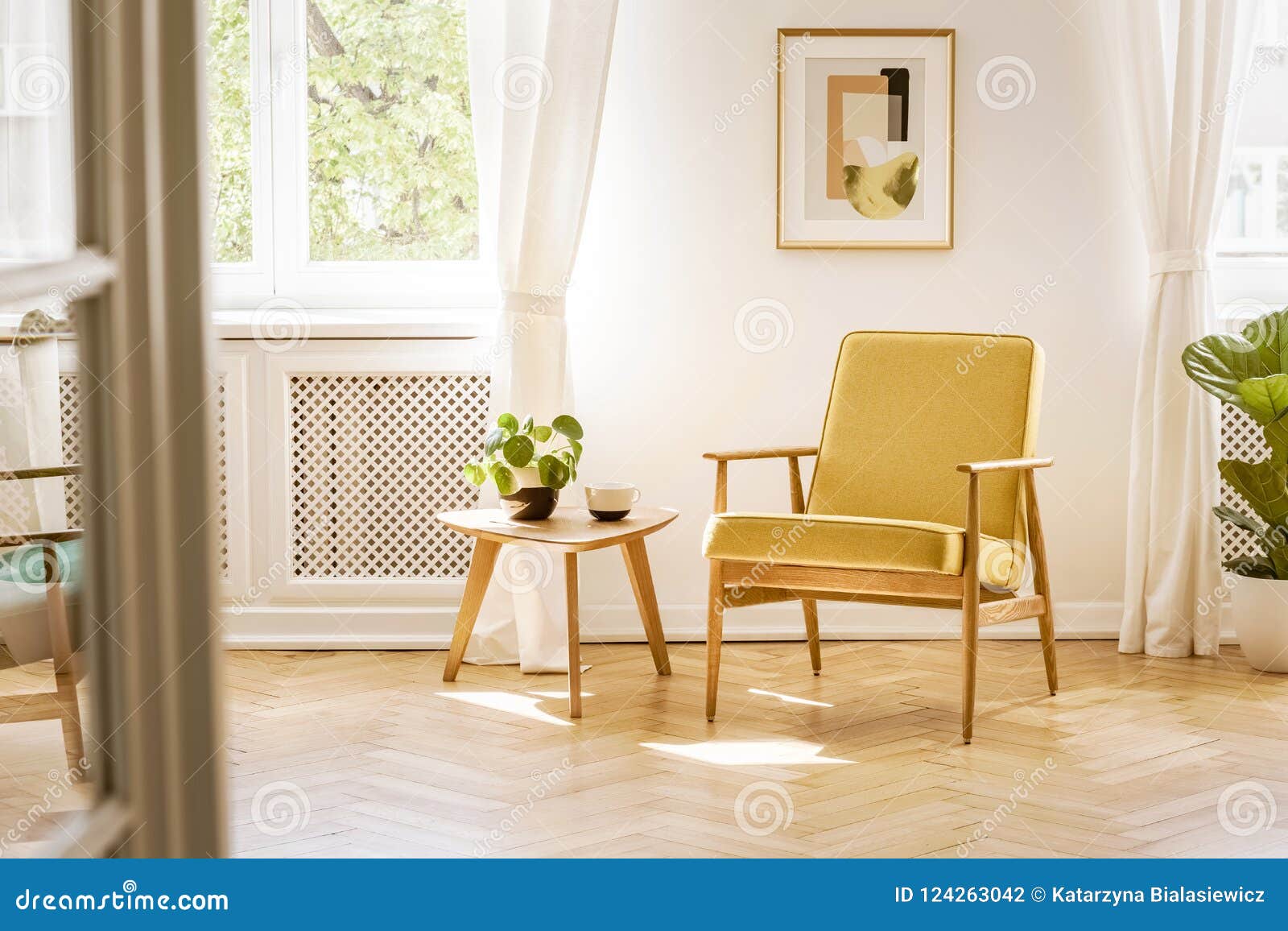 a retro, yellow armchair and a wooden table in a beautiful, sunny living room interior with herringbone floor and white walls. re