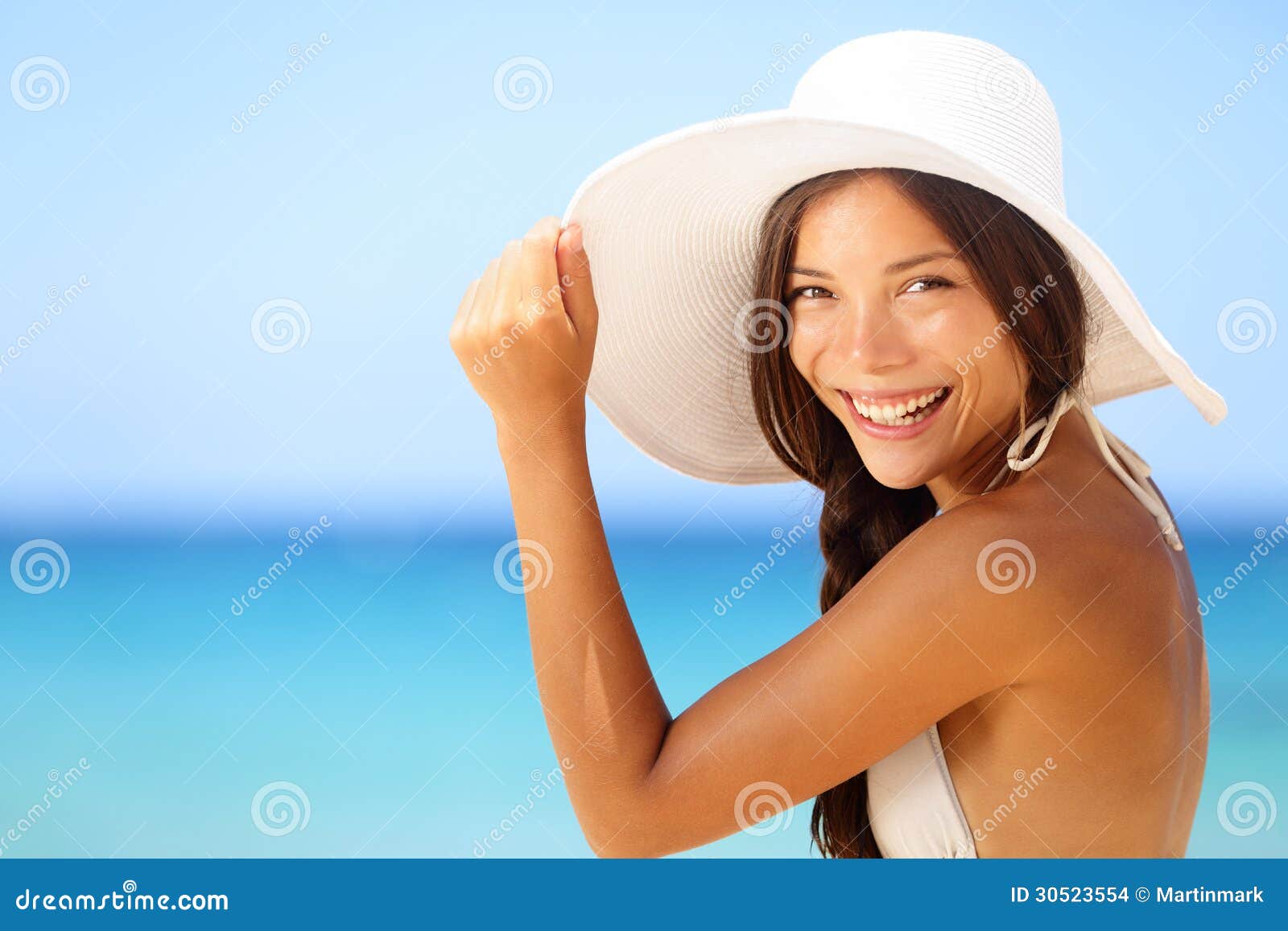 Ritratto felice sorridente della donna della spiaggia di vacanza. Ragazza asiatica del bikini sul cappello d'uso del sole della spiaggia tropicale che esamina macchina fotografica felice. Foto di stile di vita di estate con il modello femminile caucasico asiatico della corsa mista.