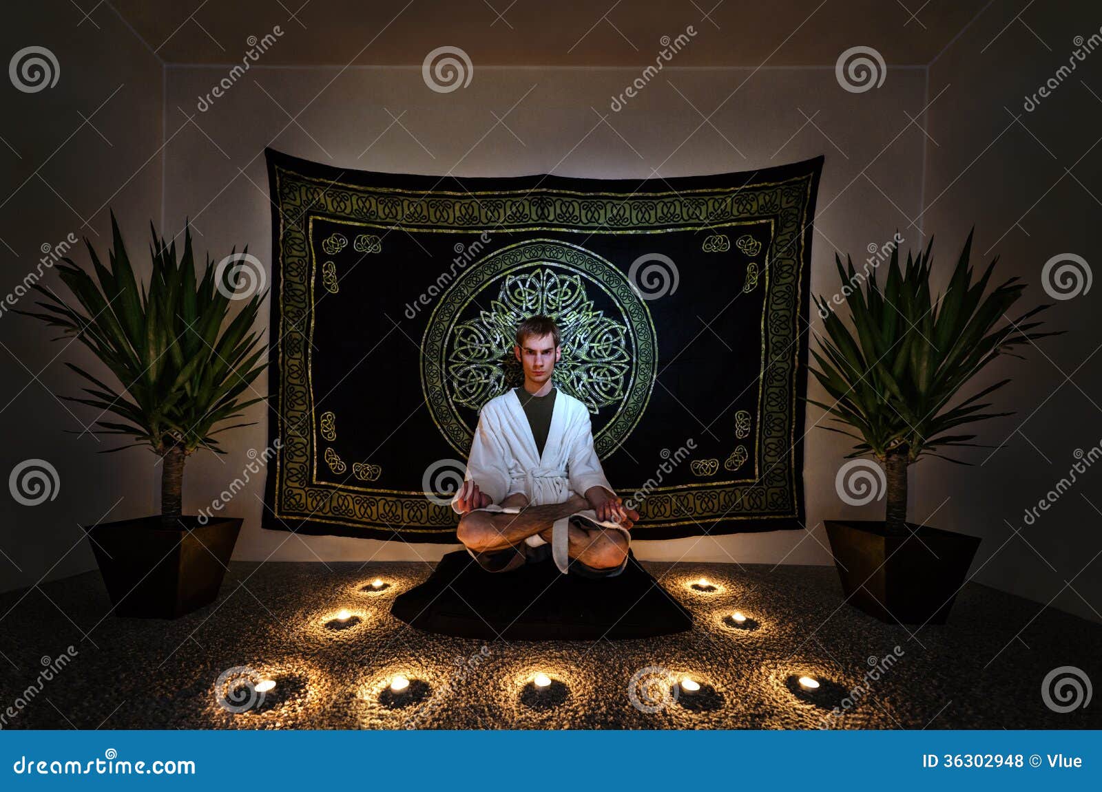 Un homme s'asseyant sur un zafu avec dedans une robe longue blanche regardant fixement intensément dans l'appareil-photo avec ses yeux ouvrent faire un rituel de méditation. Il y a des usines, des bougies, et une tapisserie derrière lui sur le mur.