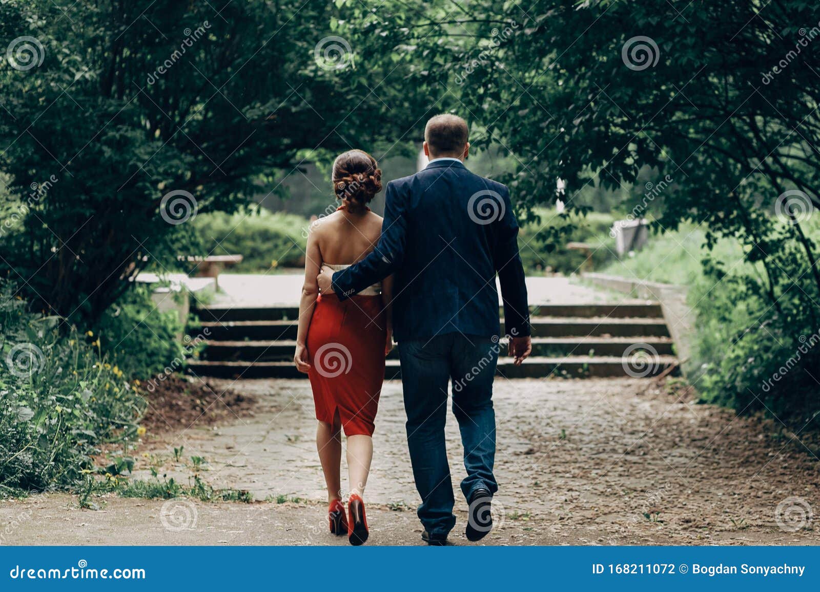 Romantic Newlywed Couple Walking In Park Handsome Goom Holding Hand Around Waist Of Bride In