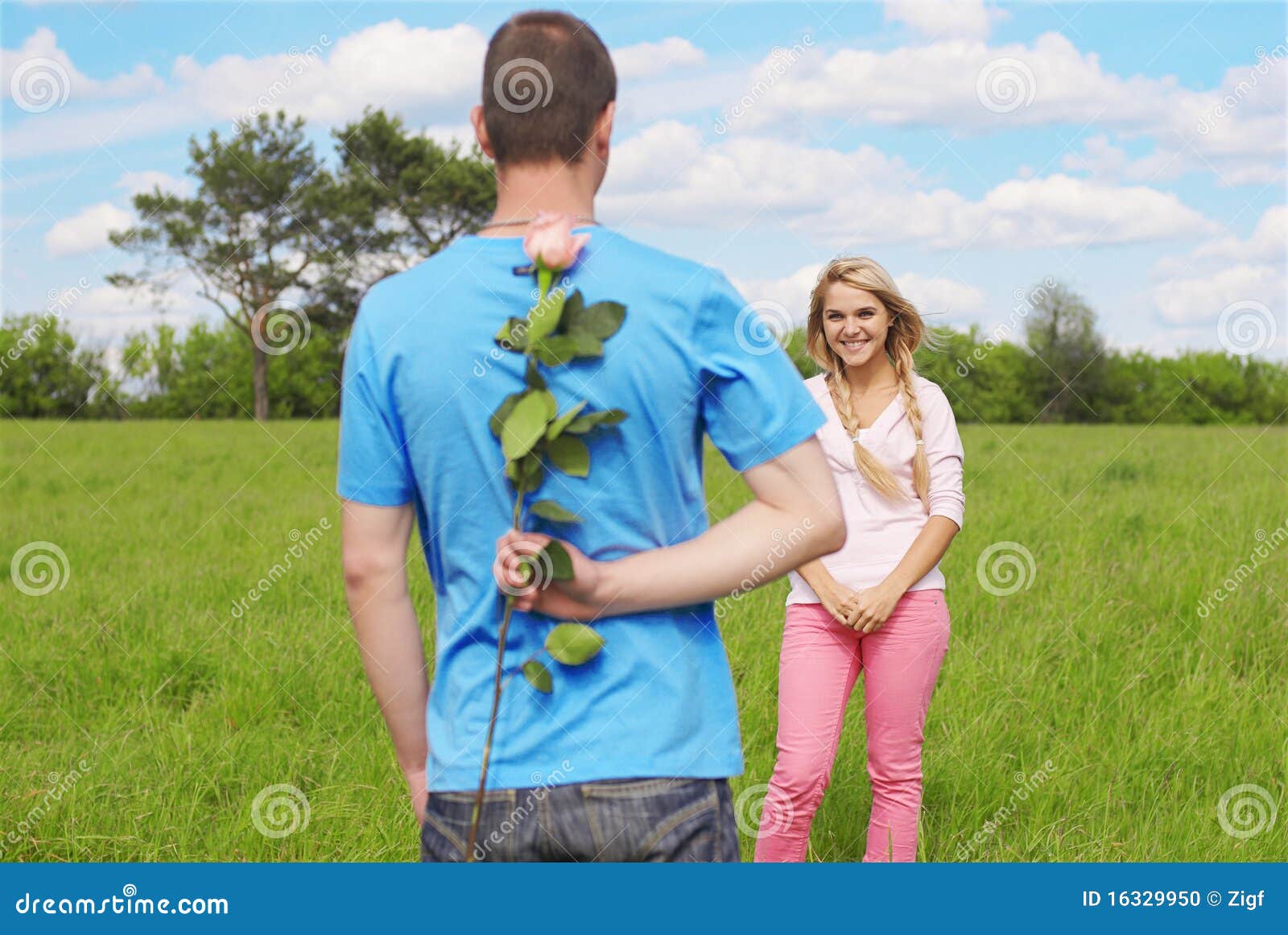 Romatic couple. Young couple outdoor on green grass. guy hiding flower beneath his back, girl looking happy. blue sky with clouds and trees on background.