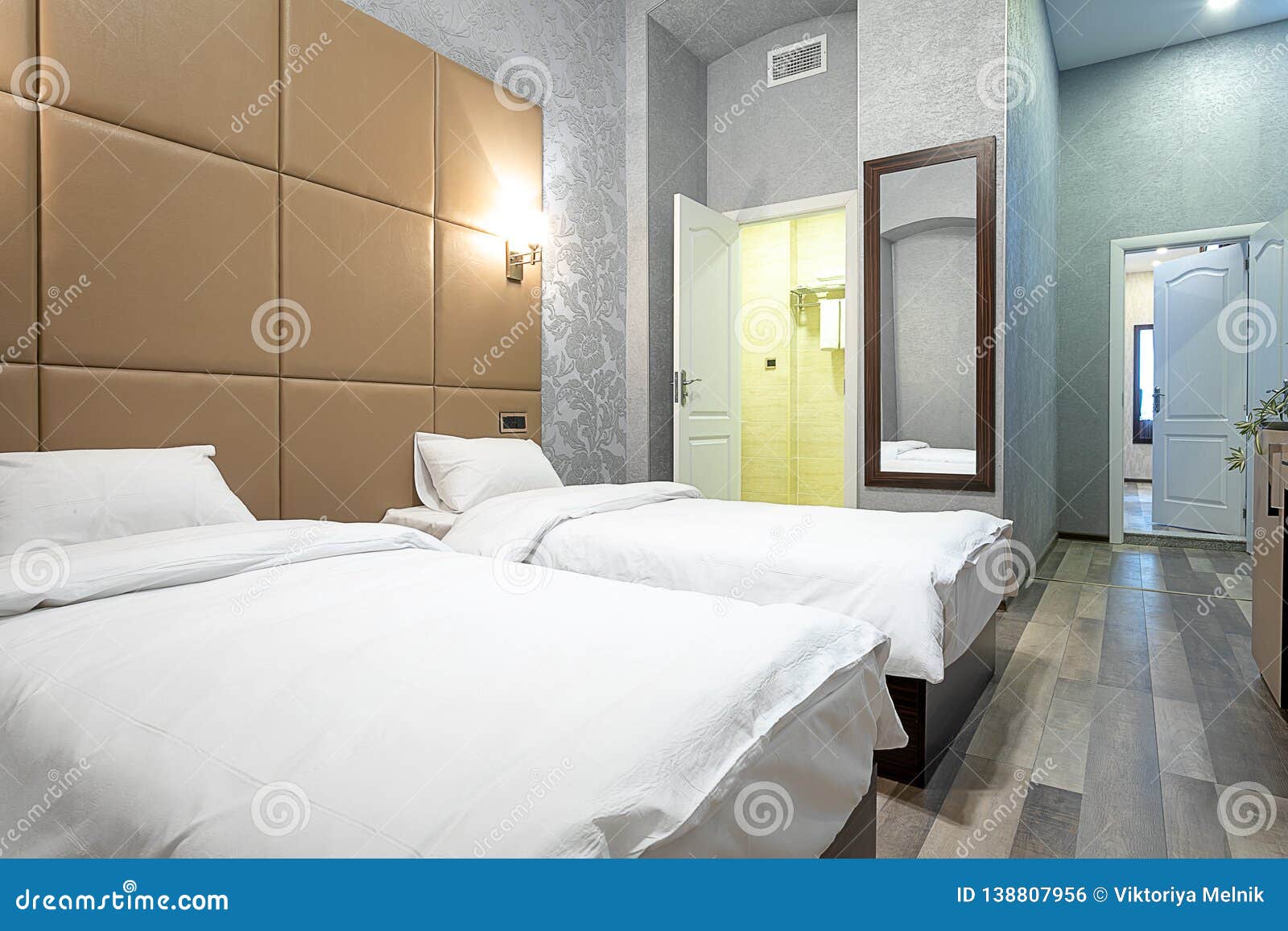 room with a double bed,bedside table,mirror on the wall,white door,light gray walls,laminate flooring and a passage to the bathroo