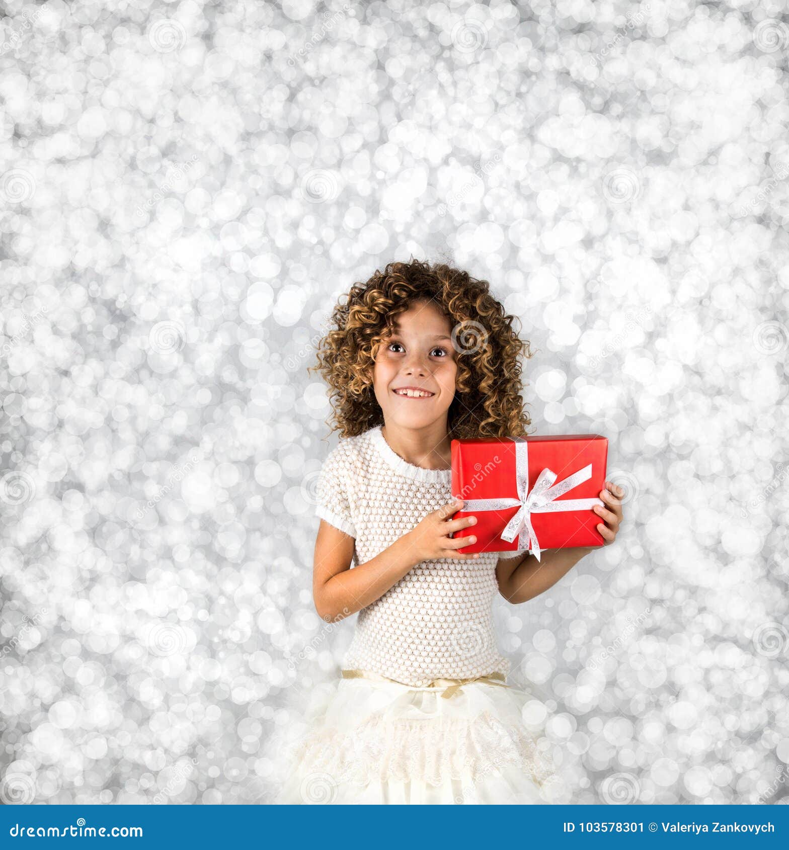 Rotes Geschenk Bild eines kleinen weißen kaukasischen Mädchens mit dem gelockten Haar, das rote Geschenkbox mit weißem Band gegen helles weißes Silber hält, beleuchtet Hintergrund