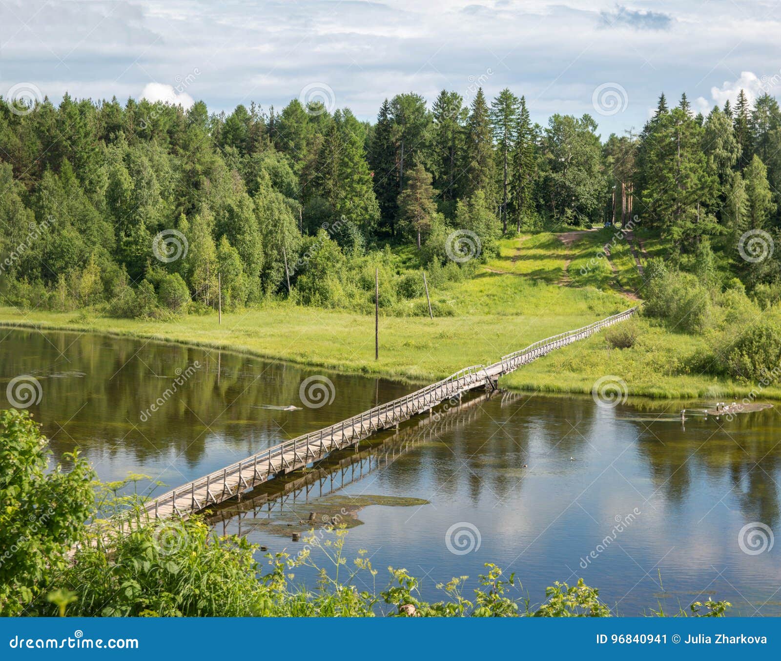 Russisches Norddorf Isady Sommertag, Emca-Fluss, alte Häuschen auf dem Ufer, alte Holzbrücke und Wolkenreflexionen