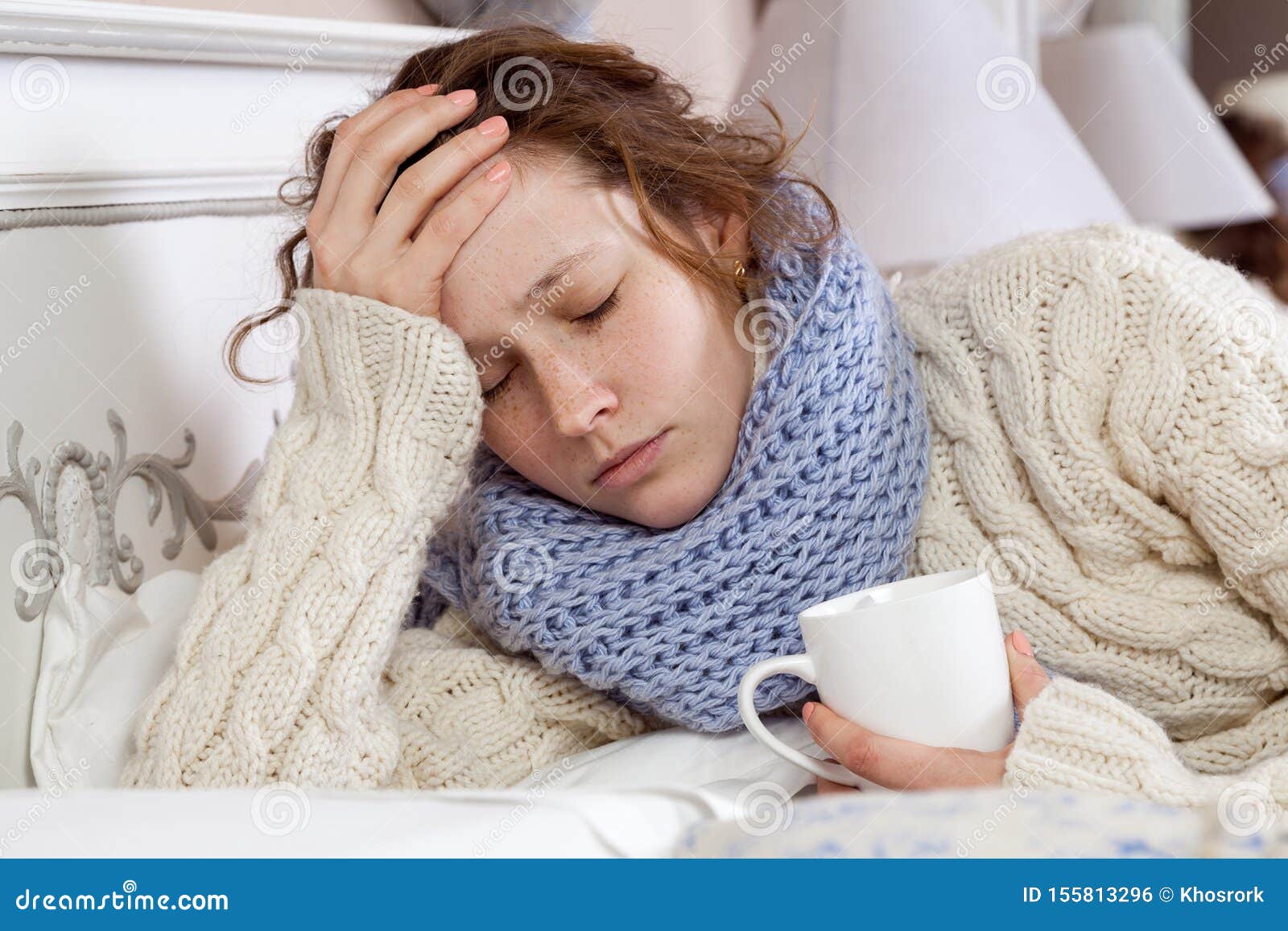 sad alone young woman in white sweater and blue scarf feeling headache, cold sick and resting home in bed. holding her painful