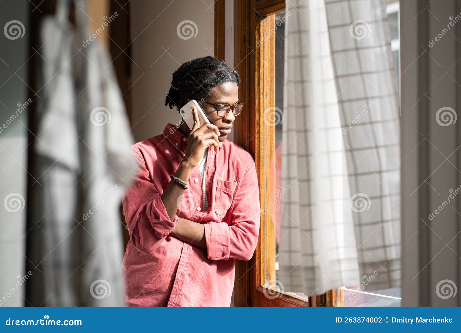 sad upset african man having difficult conversation on phone, standing by window at home