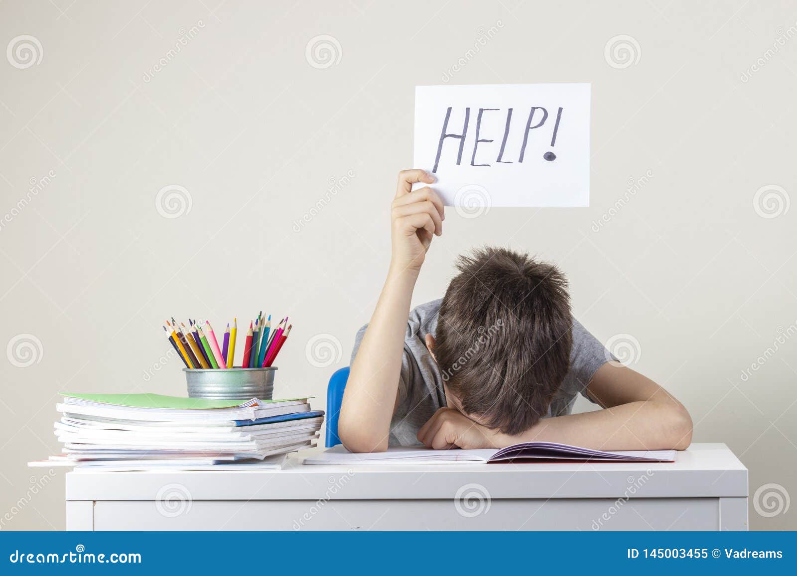 sad tired frustrated boy sitting at the table with many books and holding paper with word help. learning difficulties