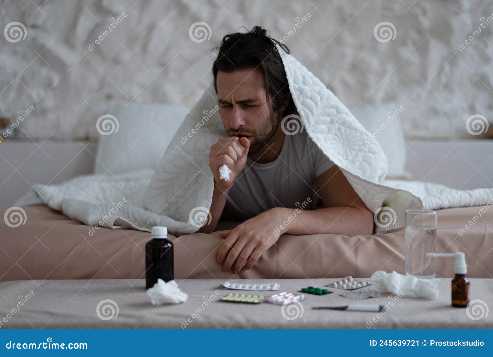 sad tired millennial european man with stubble lies under covers, coughs and treated in bedroom interior with pills