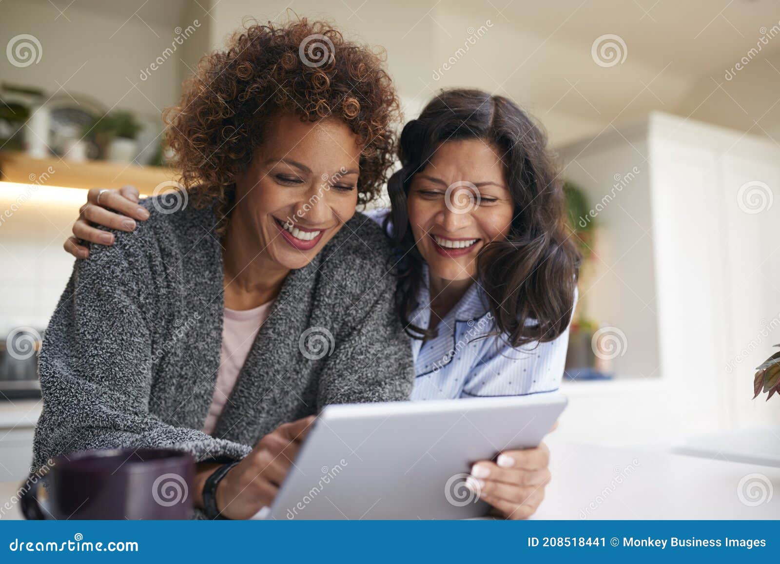 Same Sex Mature Female Couple at Home in Kitchen in Pyjamas Drinking Coffee with Digital Tablet Stock Image picture