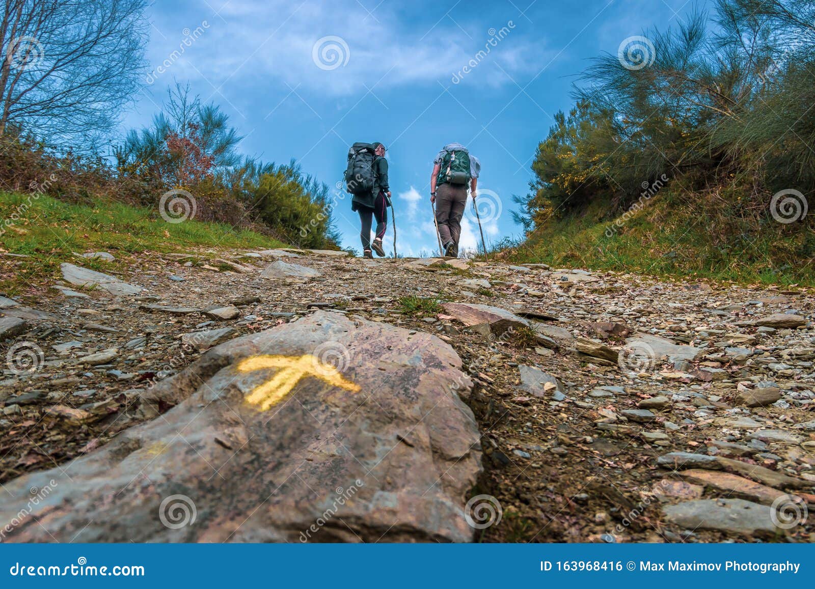 sarria, spain - two pilgrims hiking up a hill with the yellow arrow way mark outside sarria along the way of st james el camino