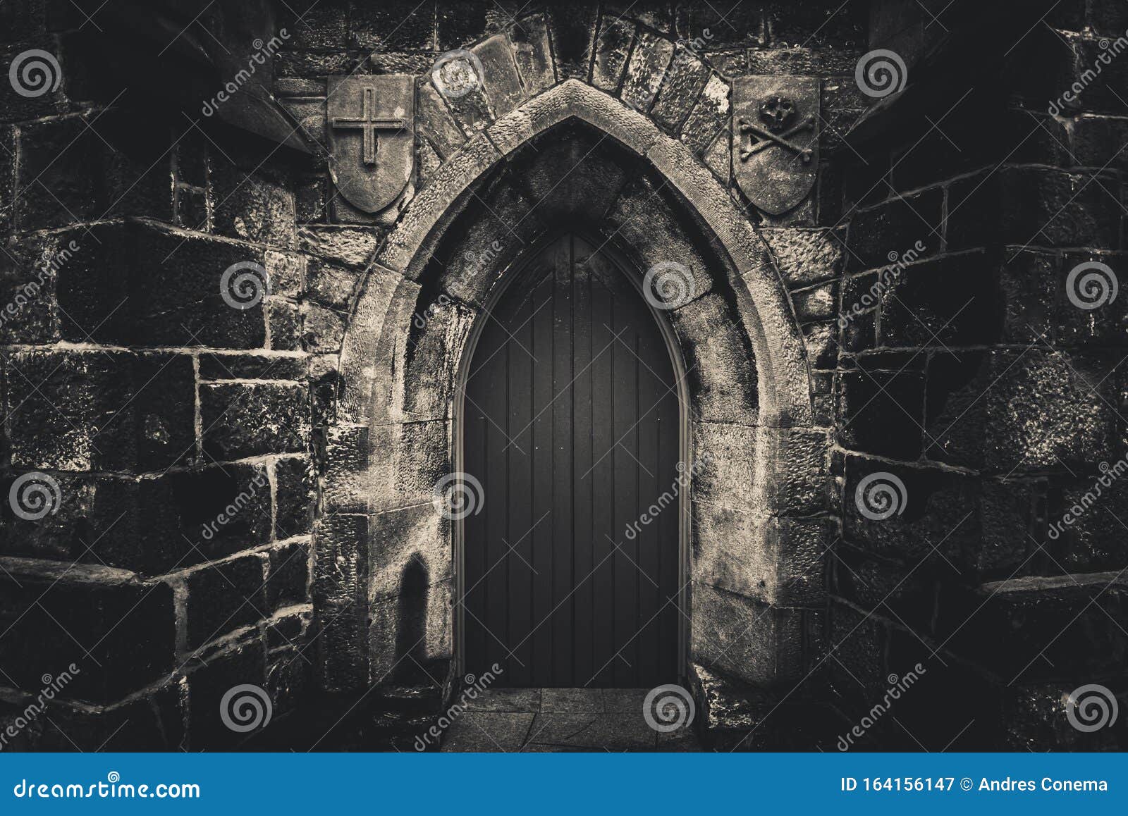 scary pointy wooden door in an old and wet stone wall building with cross, skull and bones at both sides in black and white.
