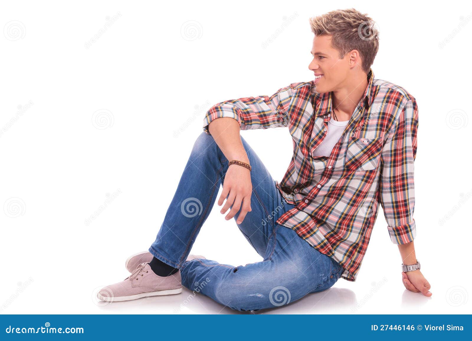 Seated casual man looking to side. Young casual man posing on the floor and smiling while looking to his side, against white background