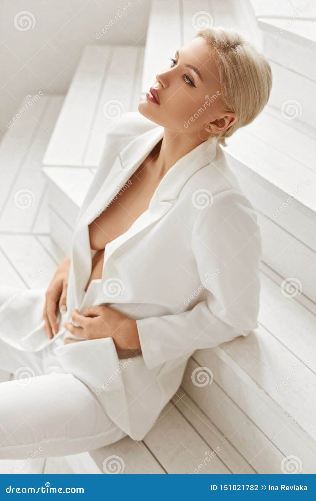 Seksowna busty młoda blondynka modela kobieta z doskonalić ciałem w garniturze na nagim ciele siedzi na bielu drewnianym. Seksowna busty młoda blondynka modela kobieta z doskonalić ciałem w garniturze na nagim ciele siedzi na białych drewnianych schodkach Gorąca dziewczyny sekretarka kusi i seksowny pozować w wnętrzu
