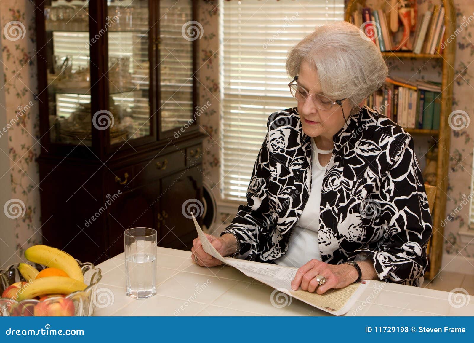 Senior Adult Woman Reading Magazine. Senior adult woman sits at counter in the kitchen with glass of water and reads magazine at home.