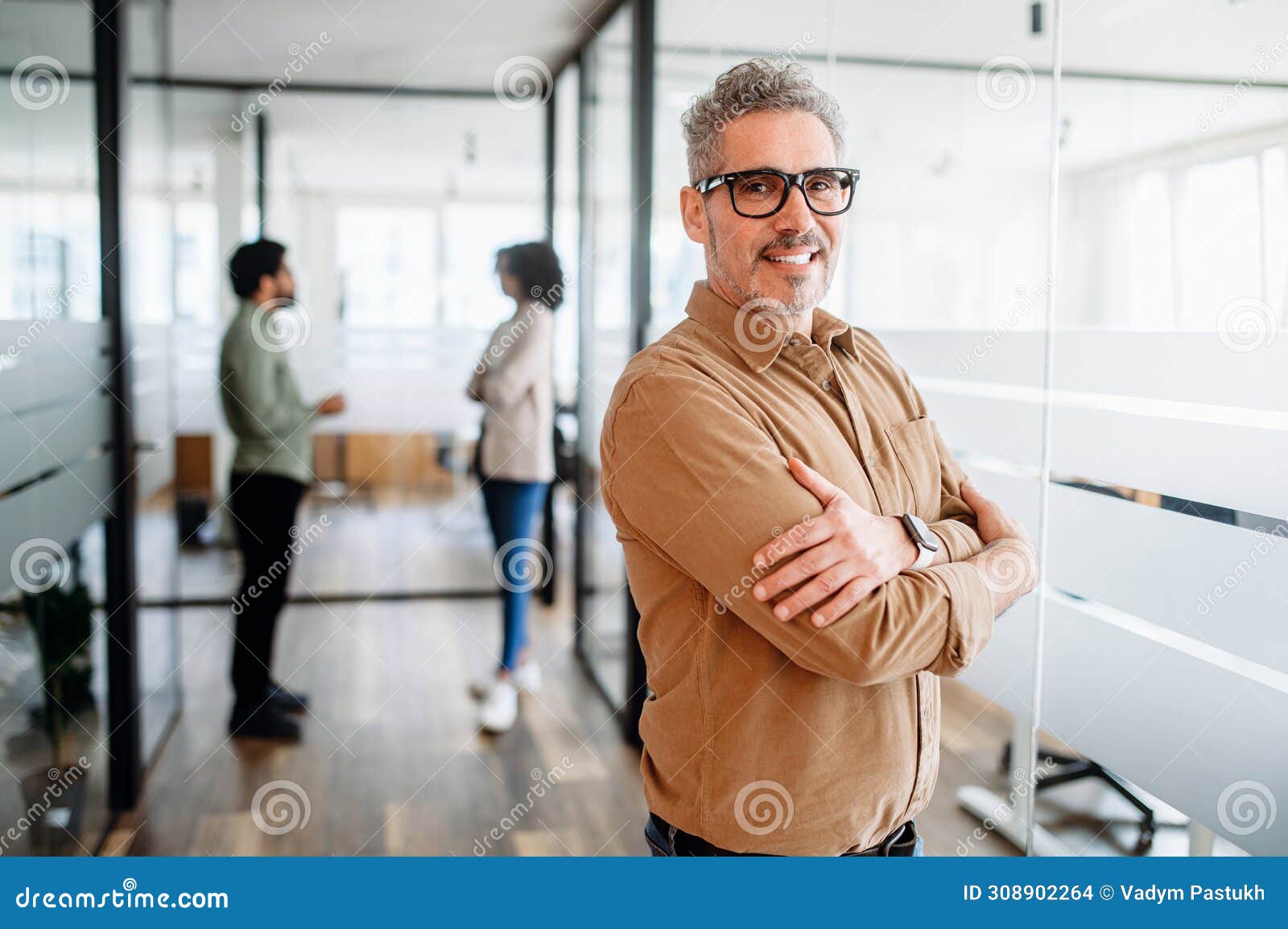 Mature man manager with a genial smile crosses his arms in satisfaction. Senior businessman in casual wear stands with crossed arms in office, a blurred conversation between colleagues in the background, a moment of individual focus within a collaborative team environment