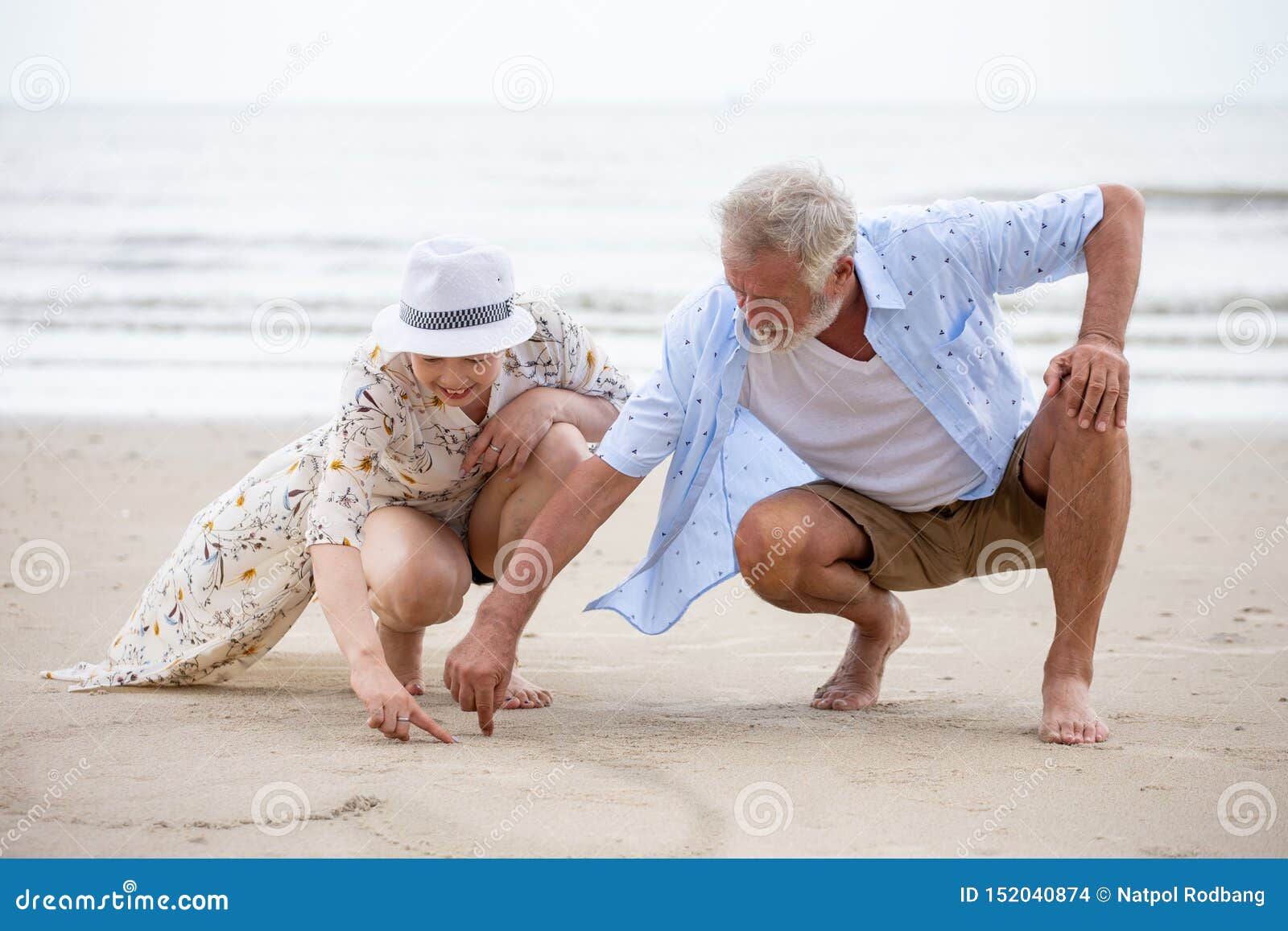 senior couple sitting on the beach drawing a heart in the sand together ,  woman asian man caucasian