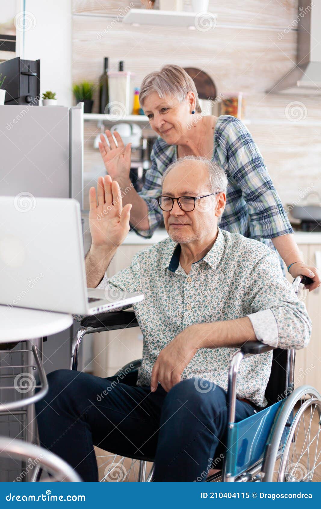 Senior Couple Waving at Webcam during a Video Call on Laptop in Kitchen. Stock Image - of white, disabled: 210404115