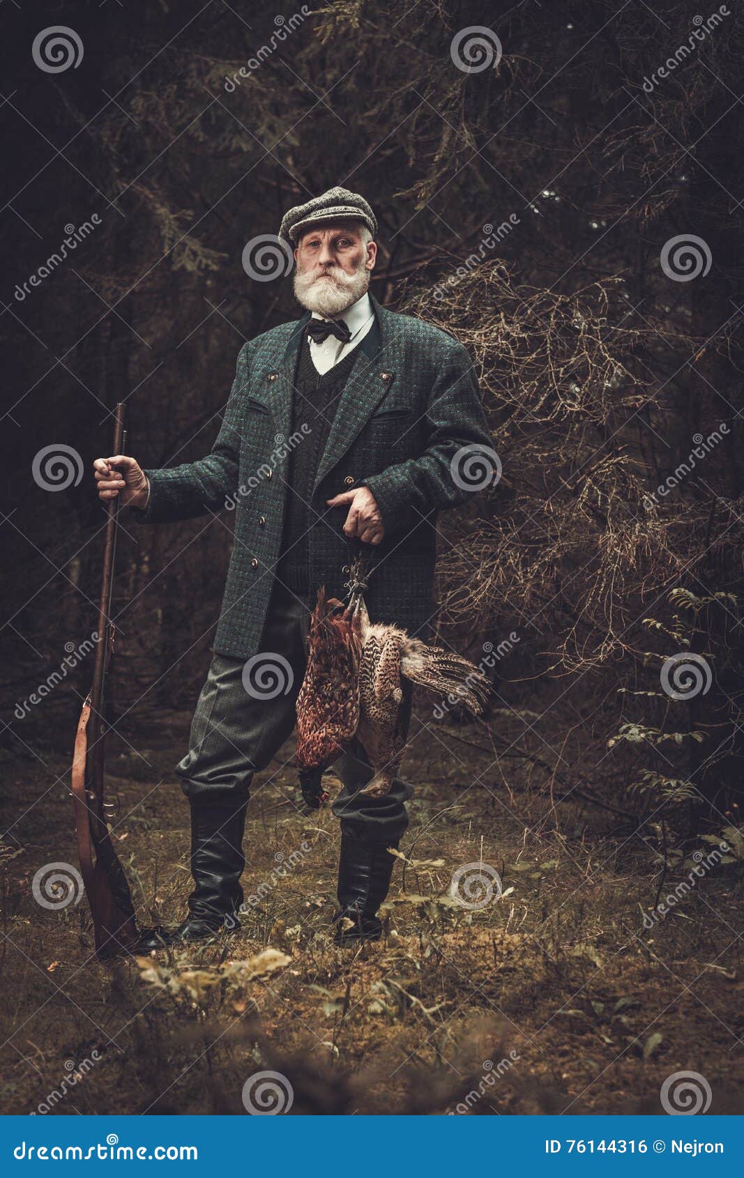 senior hunter with a shotgun and pheasants in a traditional shooting clothing, posing on a dark forest background.