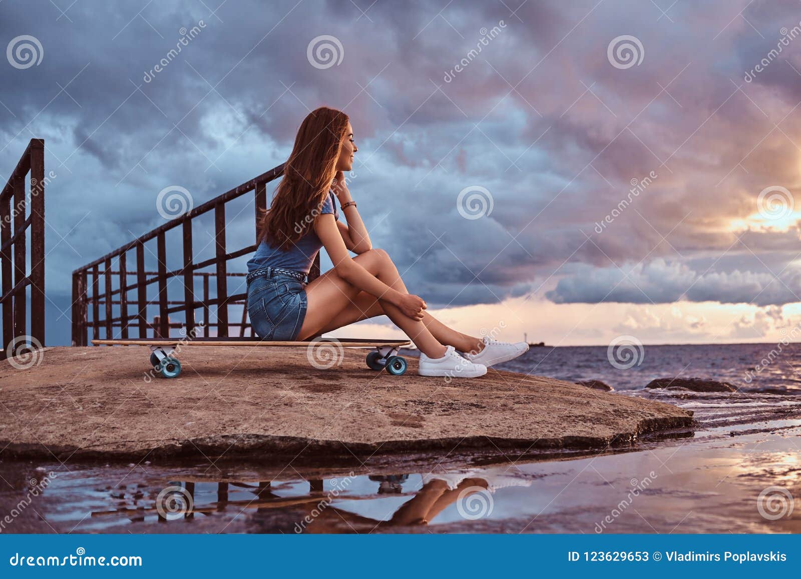 Sensual Girl Sitting On A Skateboard On The Beach Is Enjoying Amazing Dark Cloudy Weather During