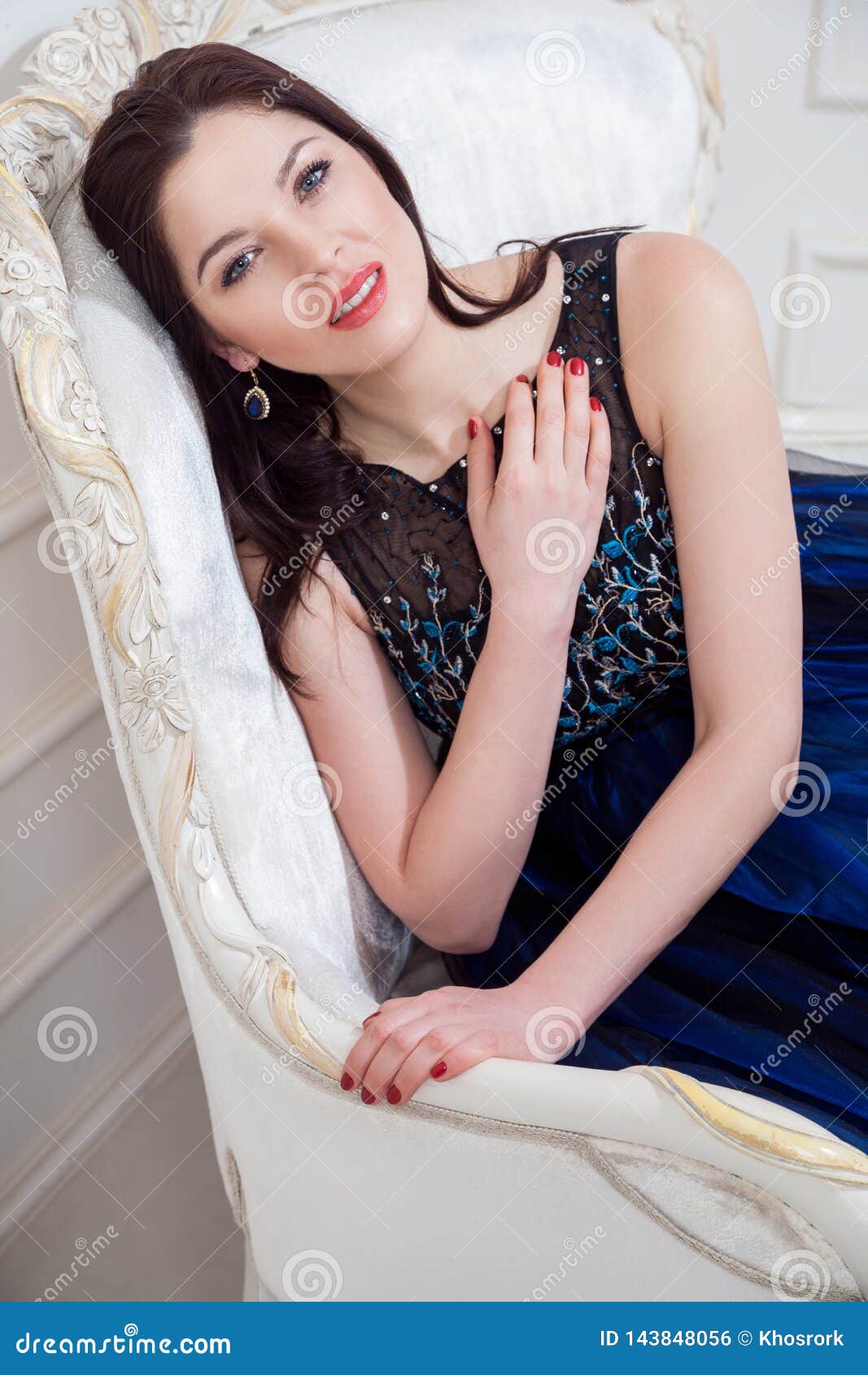 sensual woman in blue evening dress posing while sitting on vintage sofa and looking at camera with smile and pasion