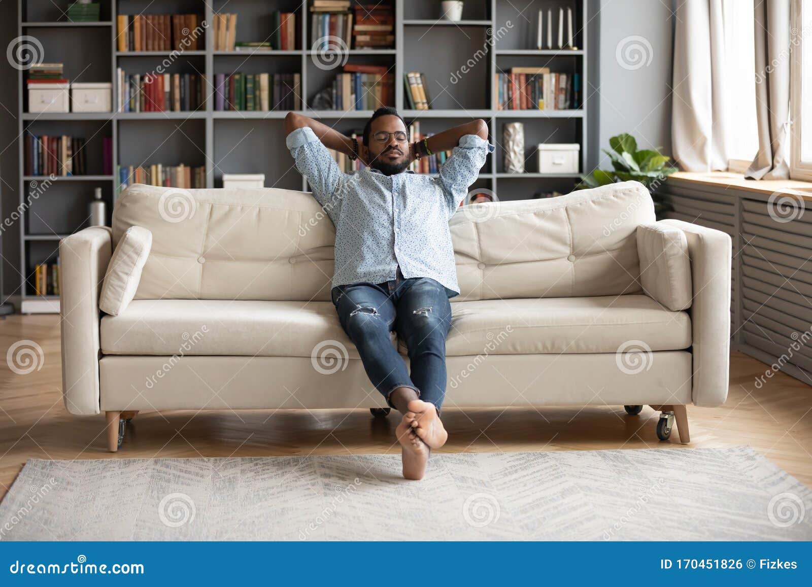 serene barefoot african man resting on sofa hands behind head