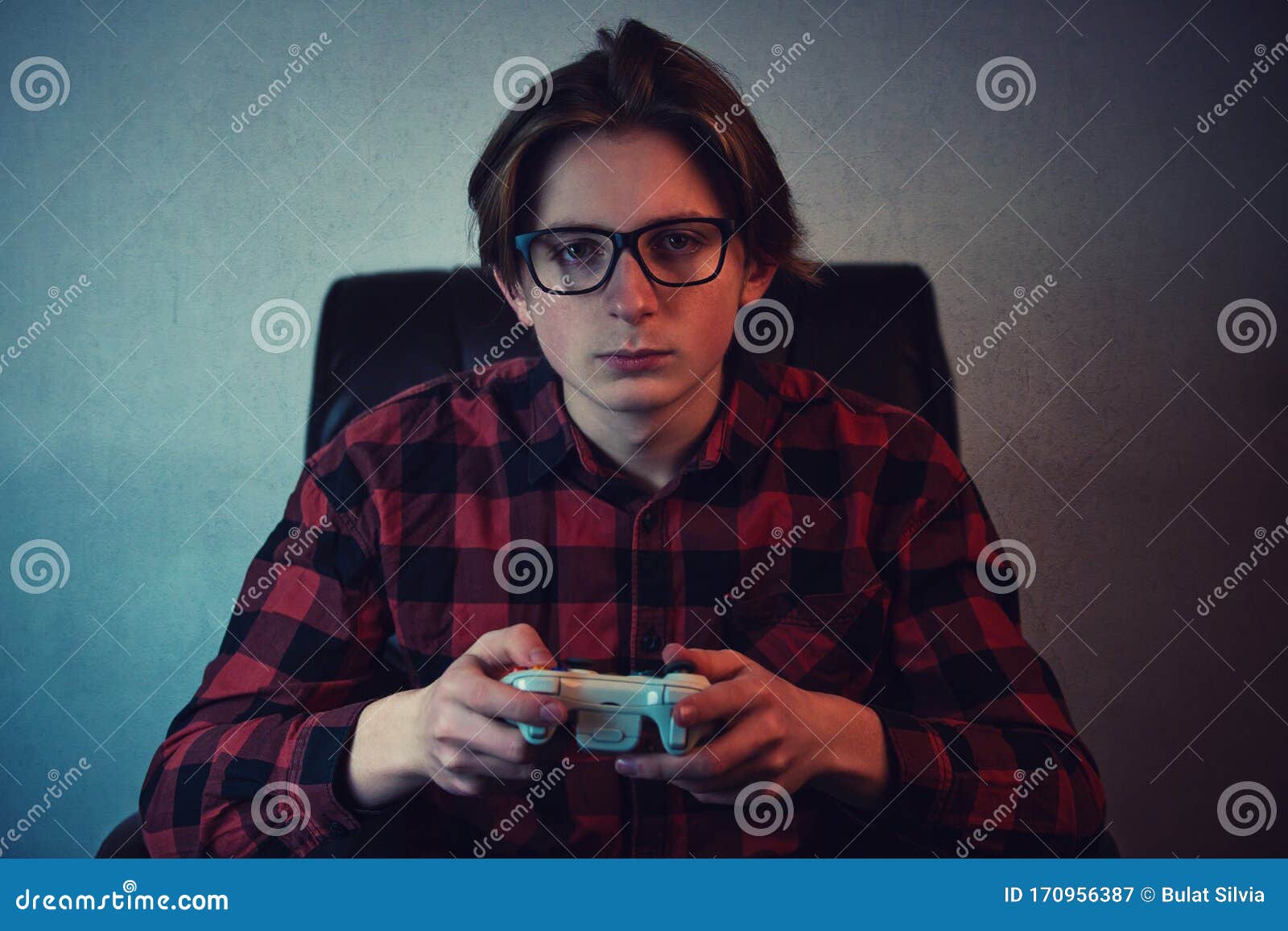 serious adolescent boy playing video games late night seated in armchair inside a dark room background. intent teen guy holding a
