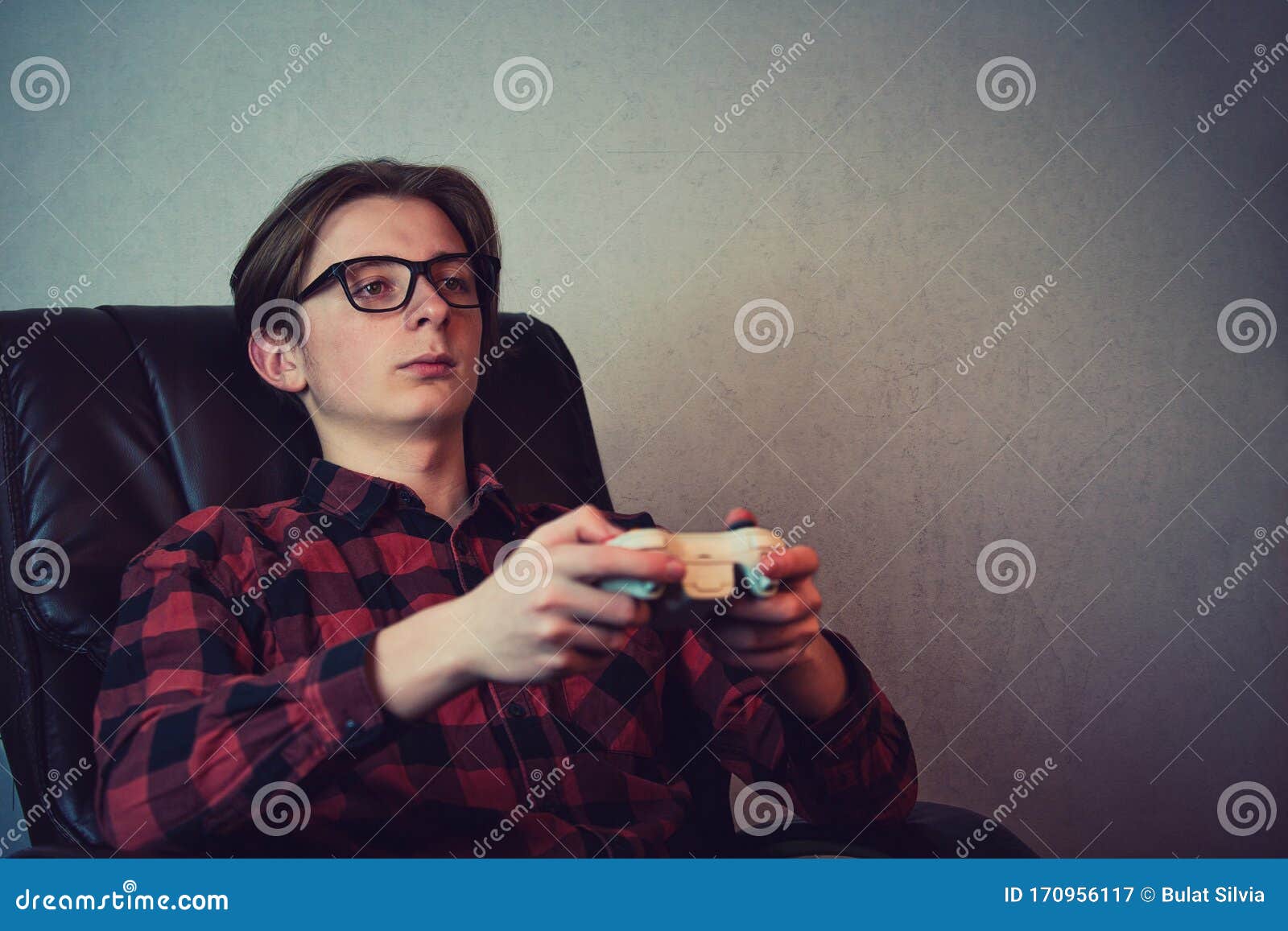 serious adolescent boy playing video games late night seated relaxed in his armchair over grey wall background. intent teen guy