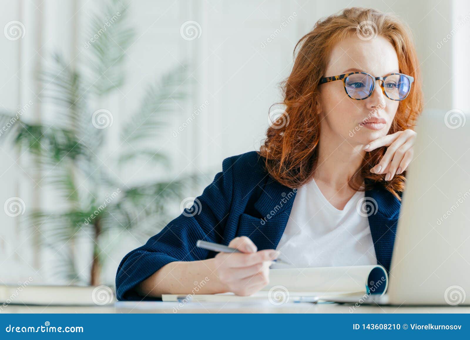 serious experienced female coach makes records in notepad, focused at screen of laptop computer, has long red hair, wears