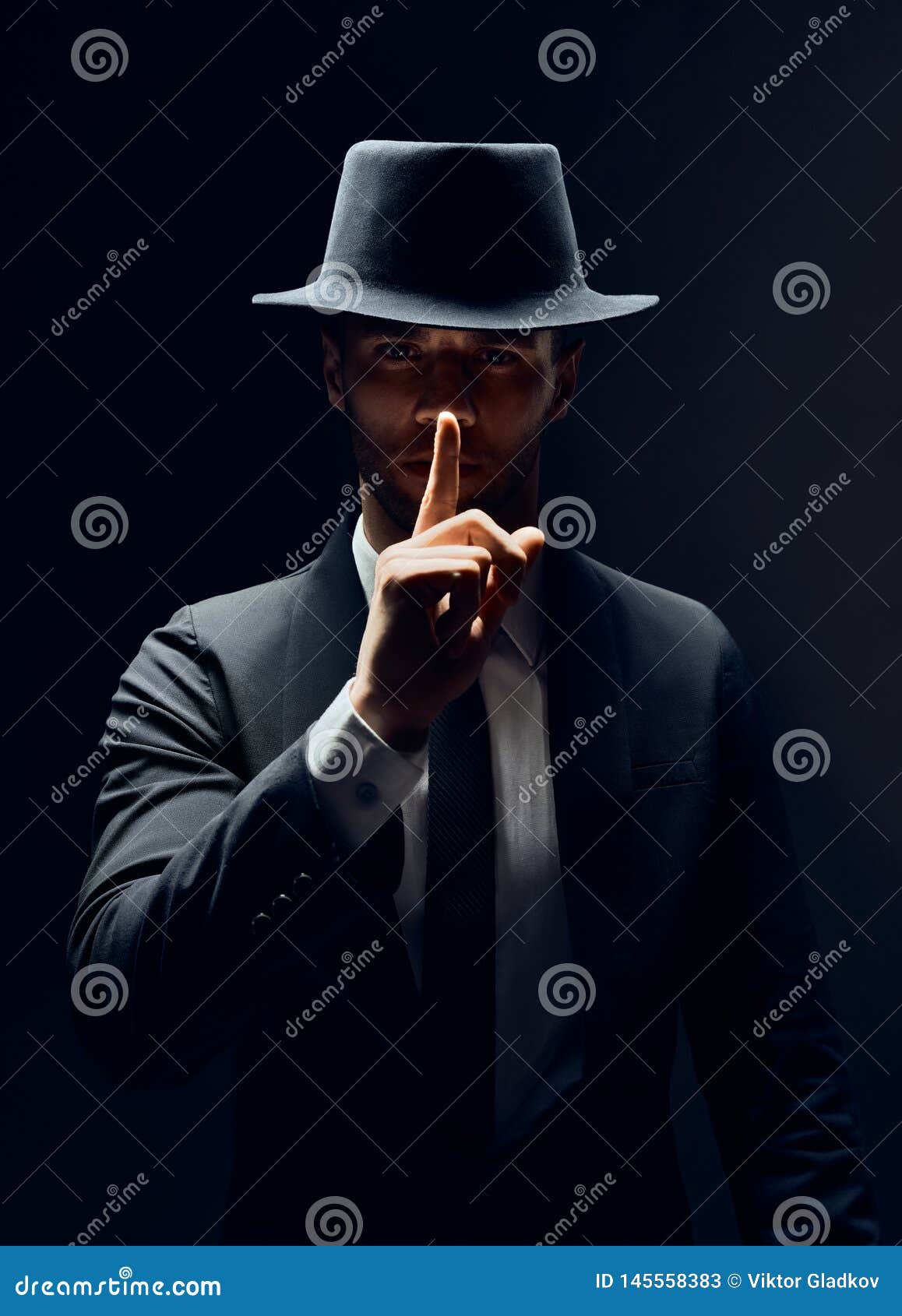 serious man in black suit and hat keeps finger on lips, making hush gesture and keep conspiracy  on dark background