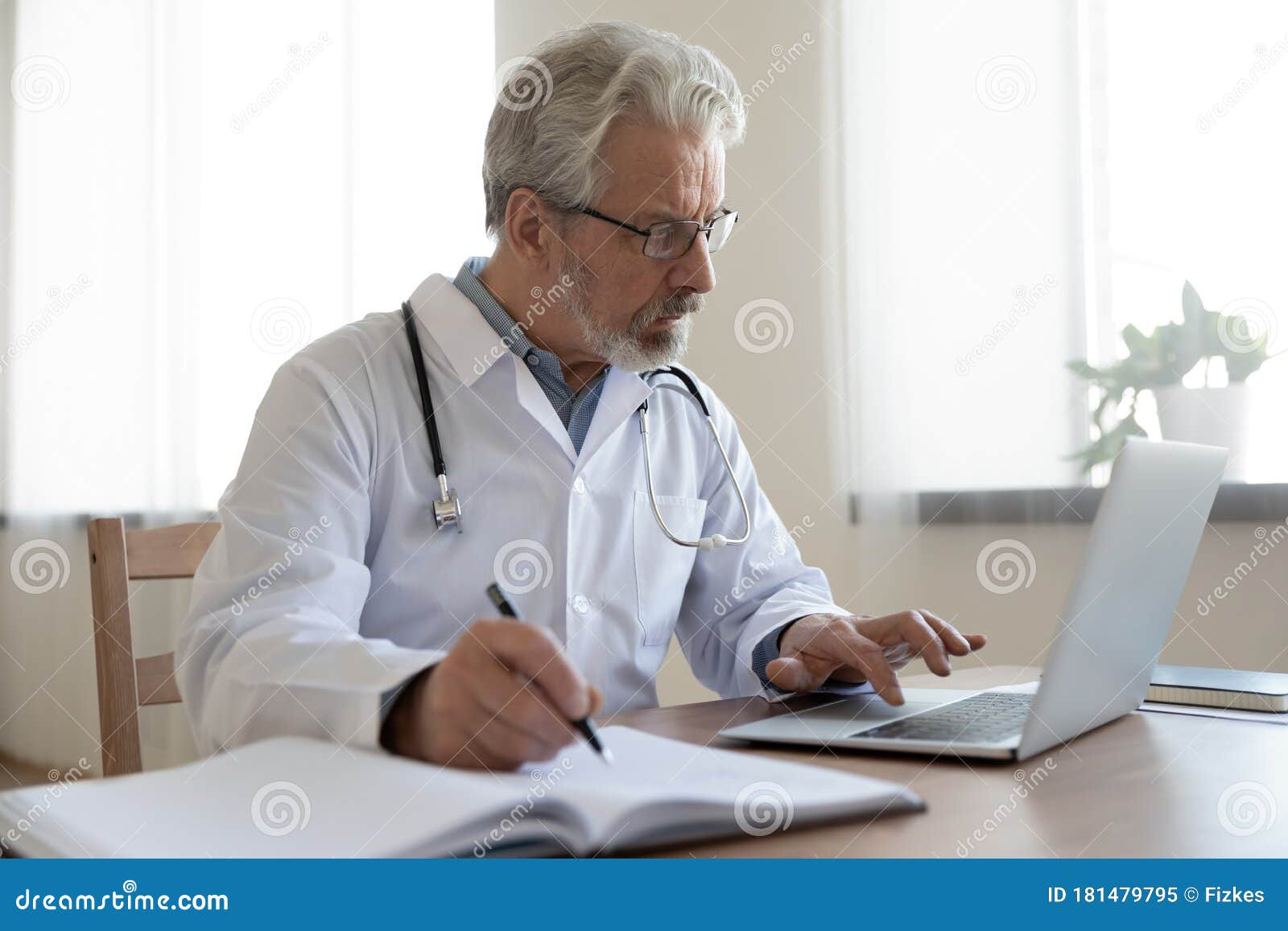 serious senior physician using computer app making notes in journal