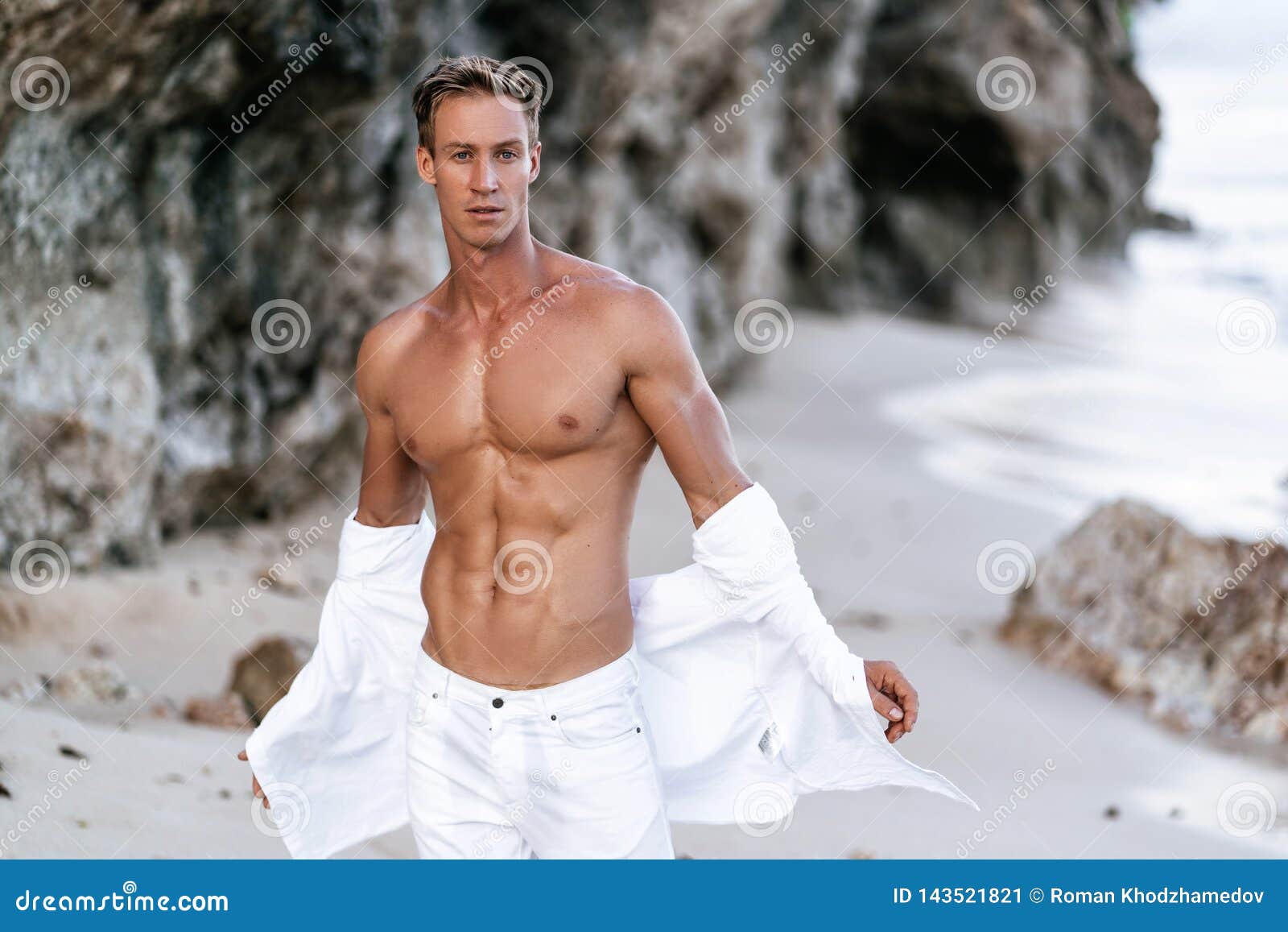 sexy muscular guy with bare-chested in white pants takes off white shirt on beach, rocks on background