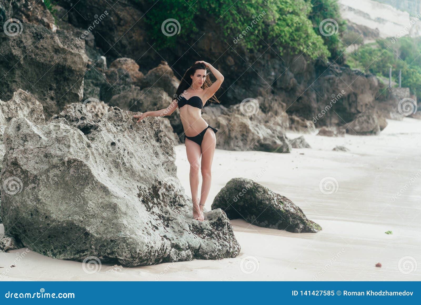 Tanned Brunette Girl In Black Swimwear Sunbathes On Beach With Rocks