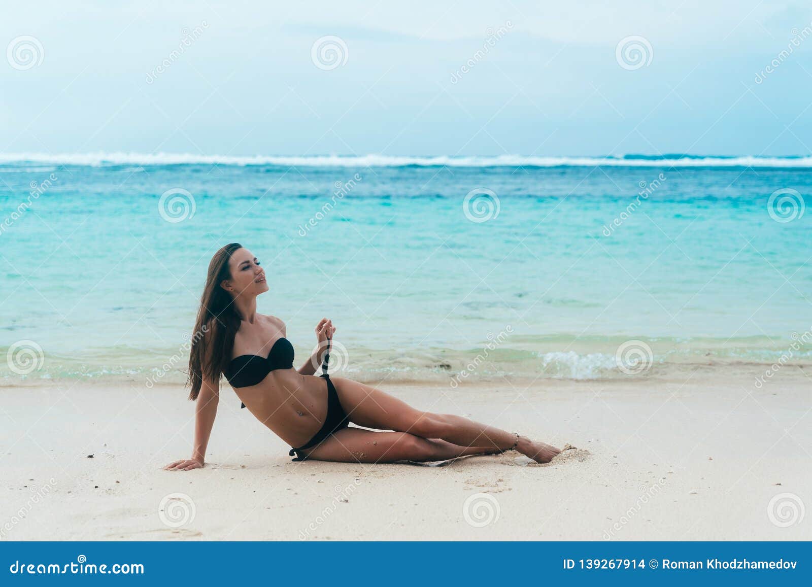 Tanned Girl In Black Swimsuit Posing On Sandy Beach Near Ocean