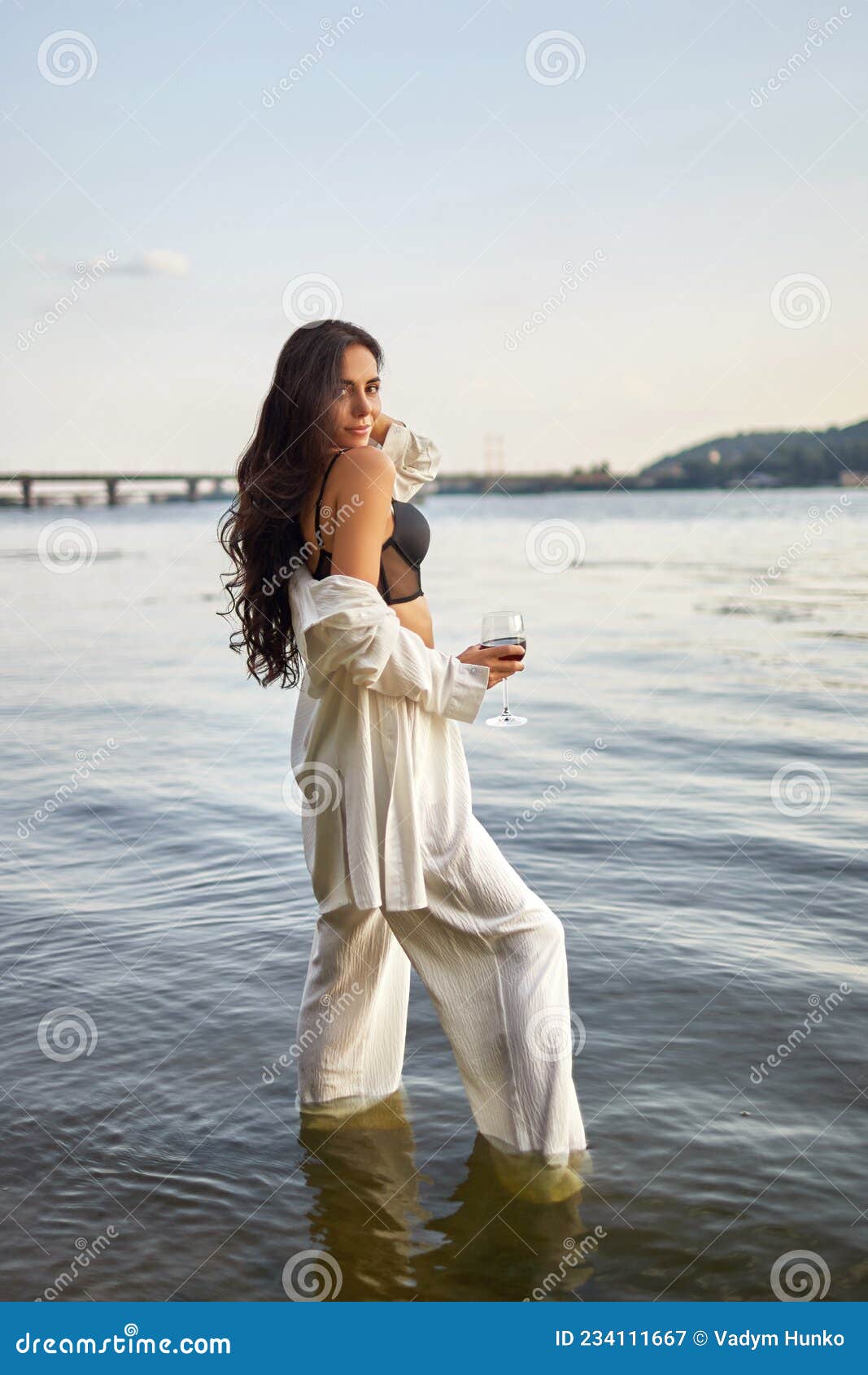 Young Brunette In A Black Bikini And White Suit With A Glass Of Wine Posing On The Beach Stock