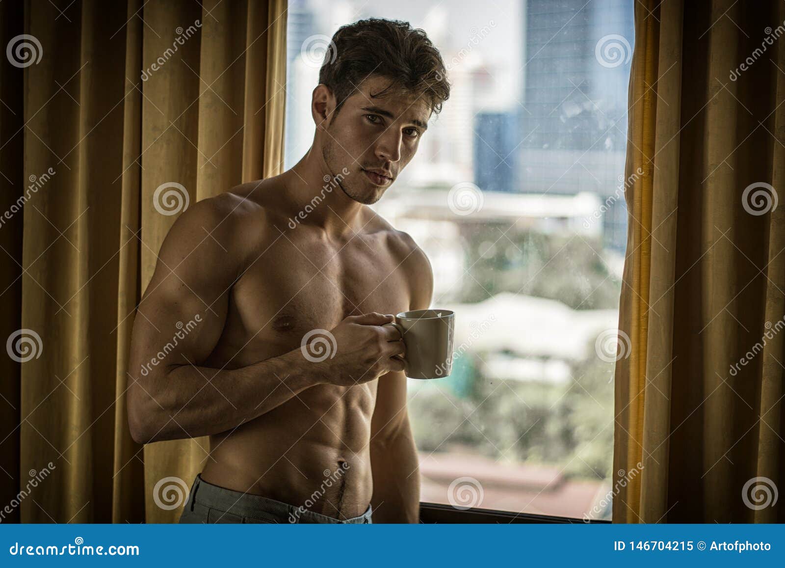 sexy young man standing shirtless by curtains with coffee